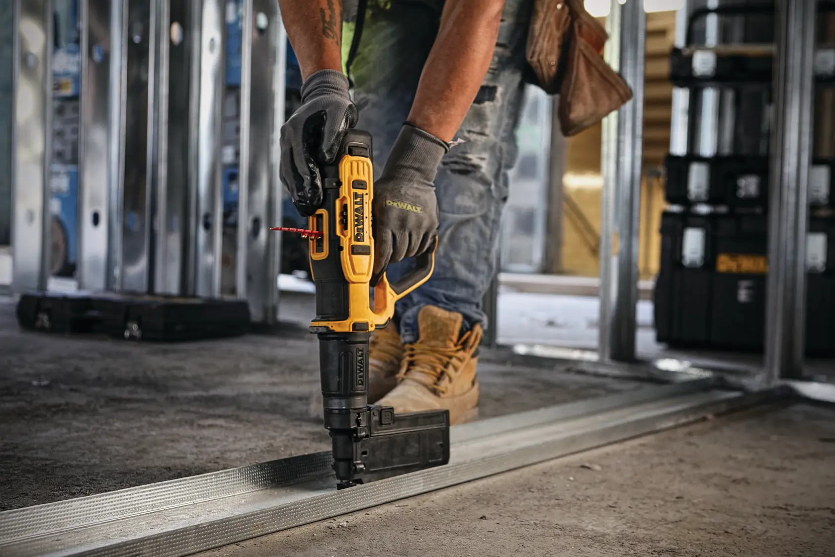 A person wearing work gloves and boots uses a DEWALT power tool to fasten metal framing on a construction site floor. The tool is held vertically against the metal, and construction materials and equipment are visible in the background.
