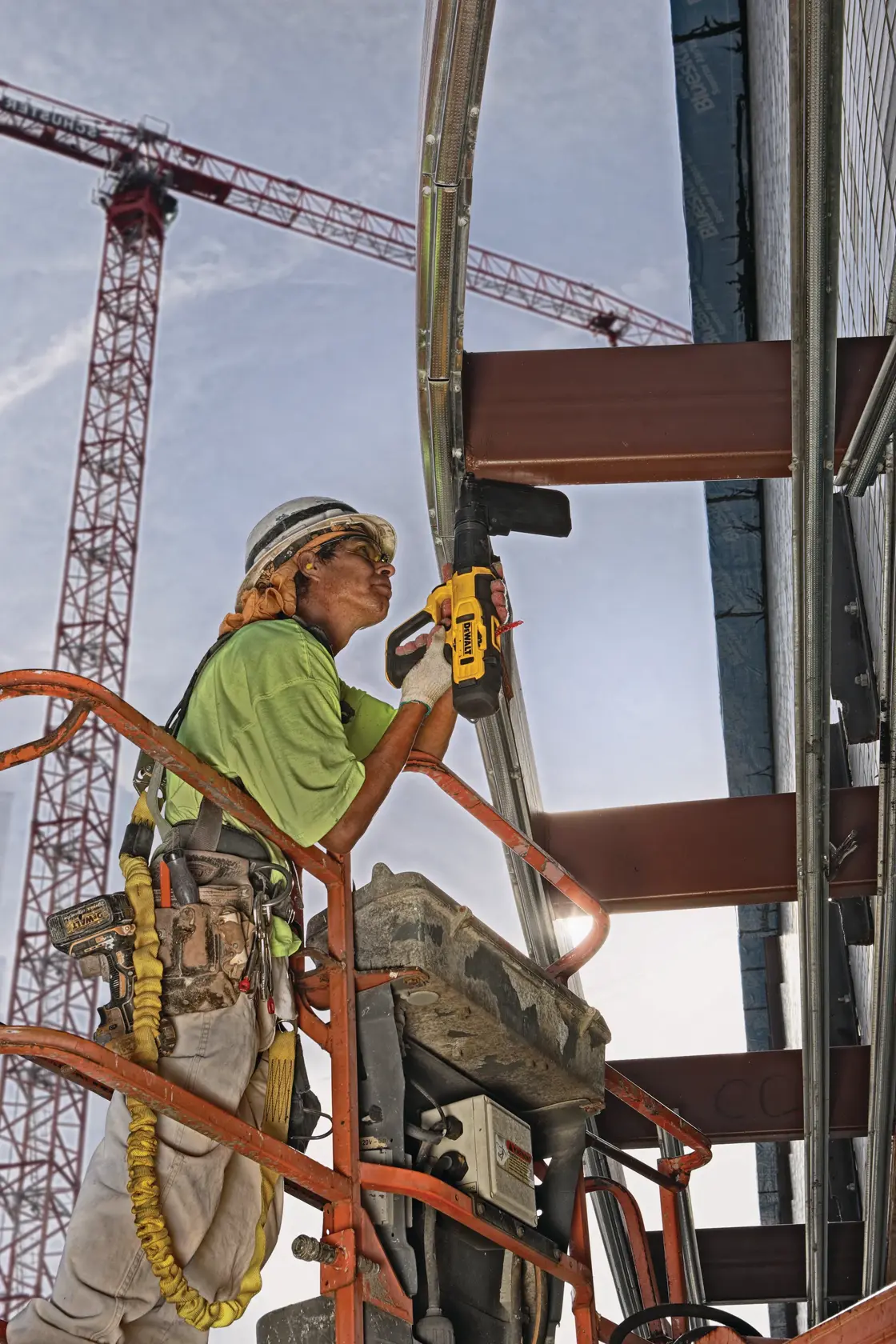 A construction worker on a lift uses a DEWALT power tool to fasten metal framing at a building site. The worker is wearing safety gear, and cranes are visible in the background.