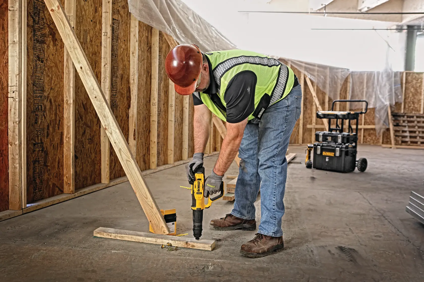 A construction worker wearing a hard hat, safety vest, jeans, gloves, and work boots is using a DEWALT power tool to fasten a wooden beam to the floor in an indoor construction site. There are unfinished wooden walls and construction equipment visible in the background.