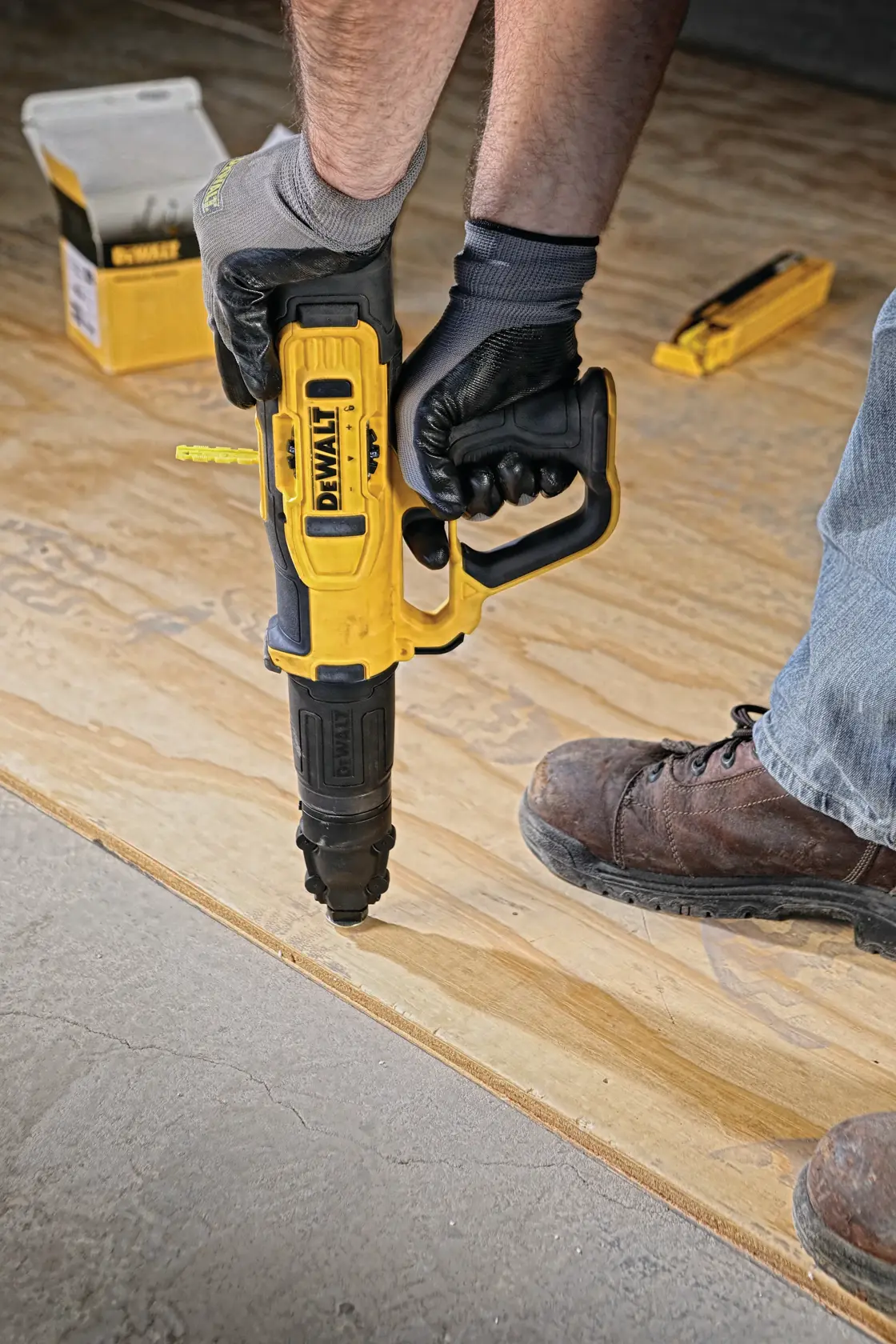 A person wearing gloves and work boots is using a DEWALT branded tool, specifically a cordless nailer, to attach plywood to a floor. Construction materials and tools are visible in the background.