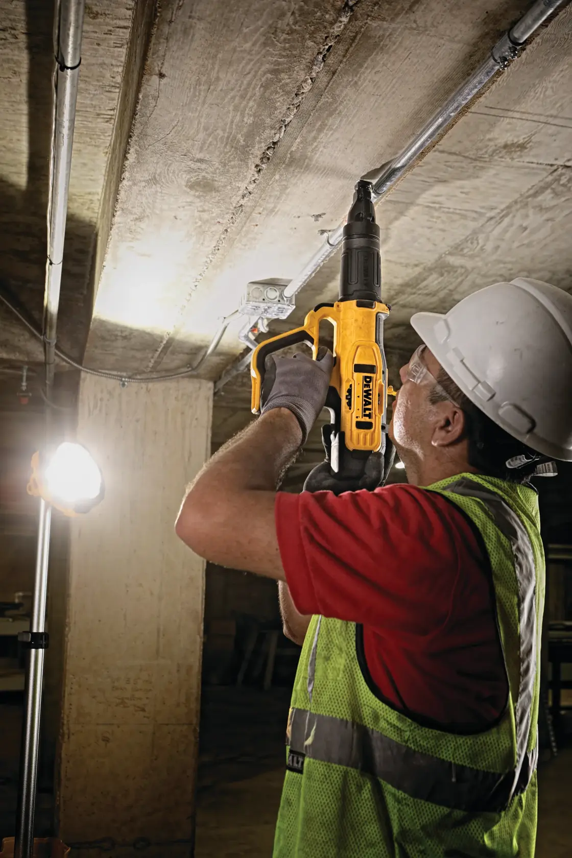 A construction worker wearing a hard hat, gloves, and reflective vest uses a yellow DEWALT tool to fasten a metal fixture to a concrete ceiling in an industrial environment.