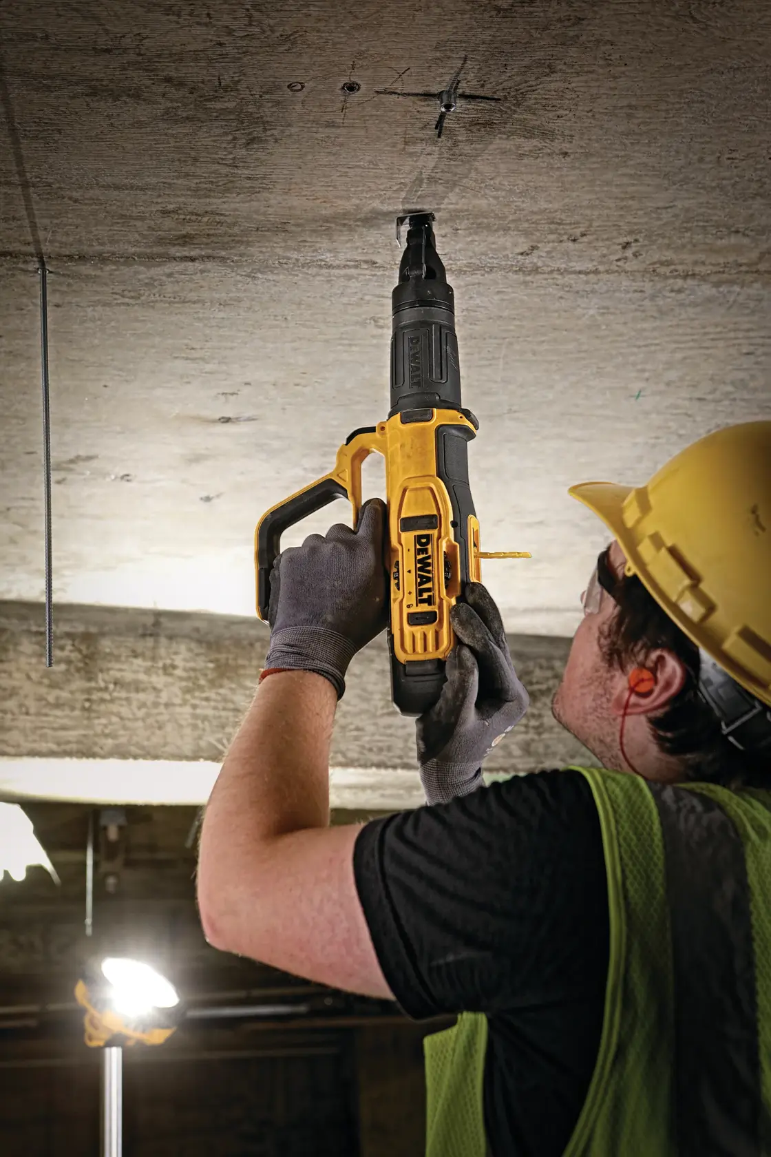 A person wearing a hard hat, safety glasses, gloves, and a green safety vest is using a yellow DEWALT power tool to drill into a concrete ceiling on a construction site.