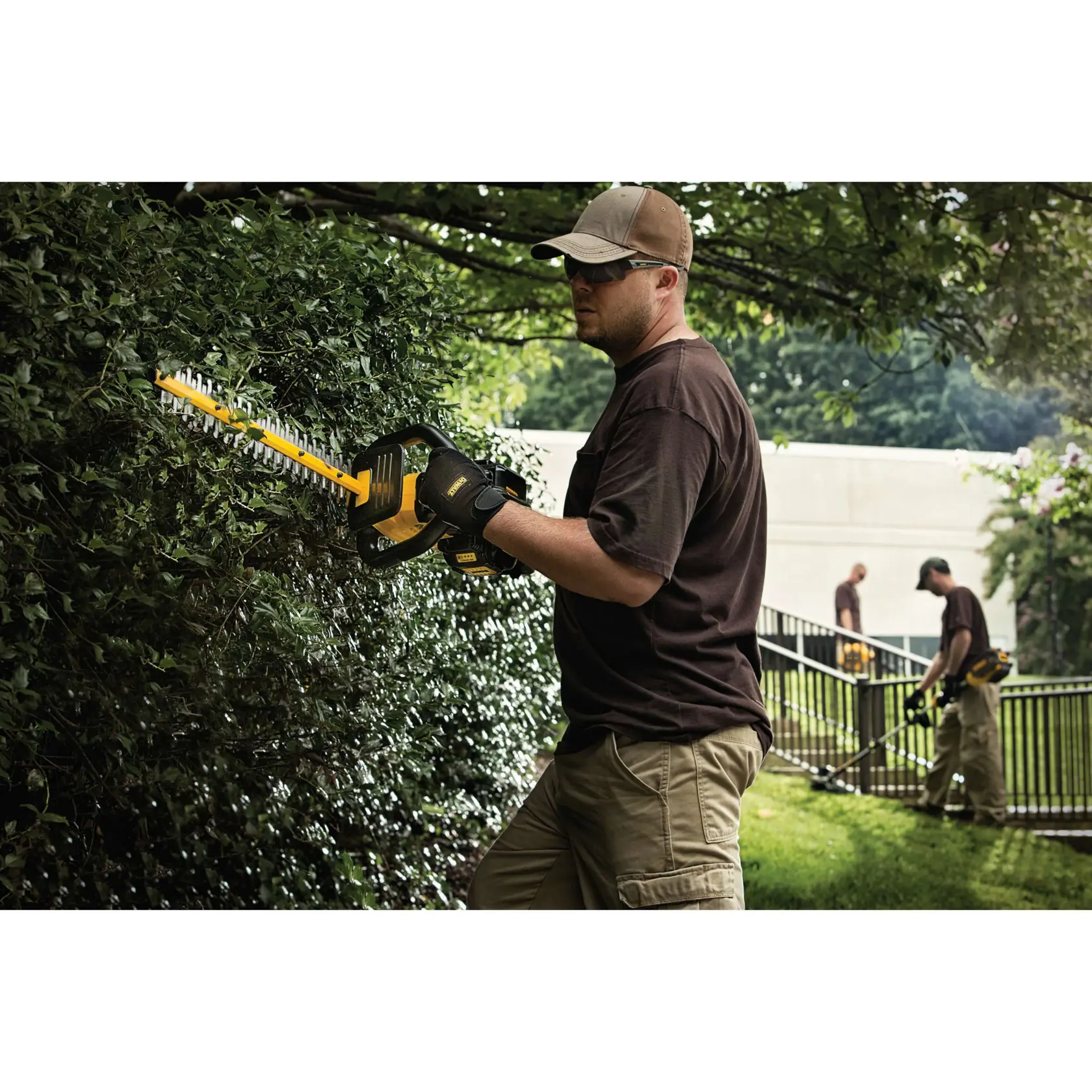 A person wearing gloves and a cap uses a DEWALT hedge trimmer to trim bushes, with two other people in the background maintaining greenery. The scene shows landscaping work outdoors.