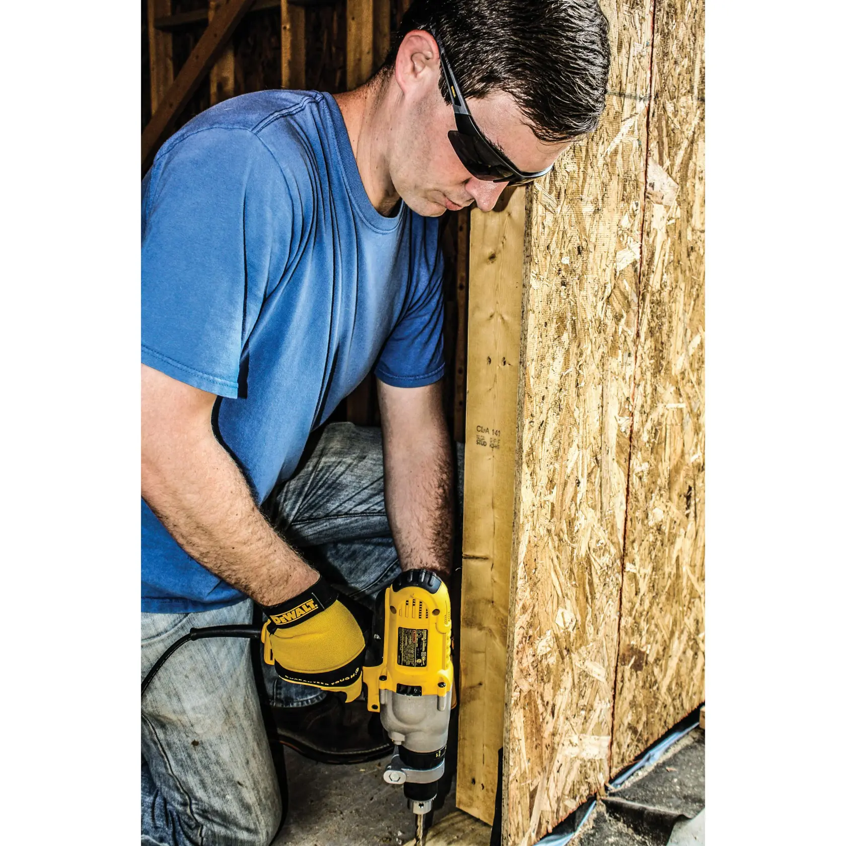 Person with their face blurred wearing a blue shirt and DEWALT-branded yellow gloves operating a yellow DEWALT power tool against a wooden structure, in a construction setting.