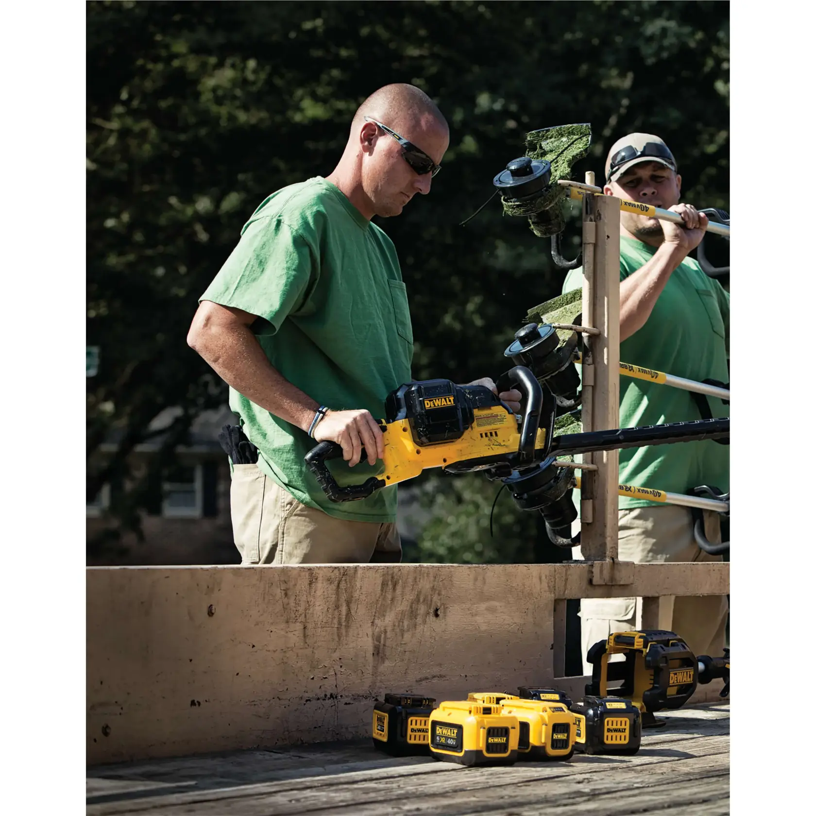 Two people wearing green shirts are using DEWALT outdoor power tools, including a hedge trimmer and string trimmer, with several DEWALT batteries placed on a wooden surface outdoors. Their faces are obscured.