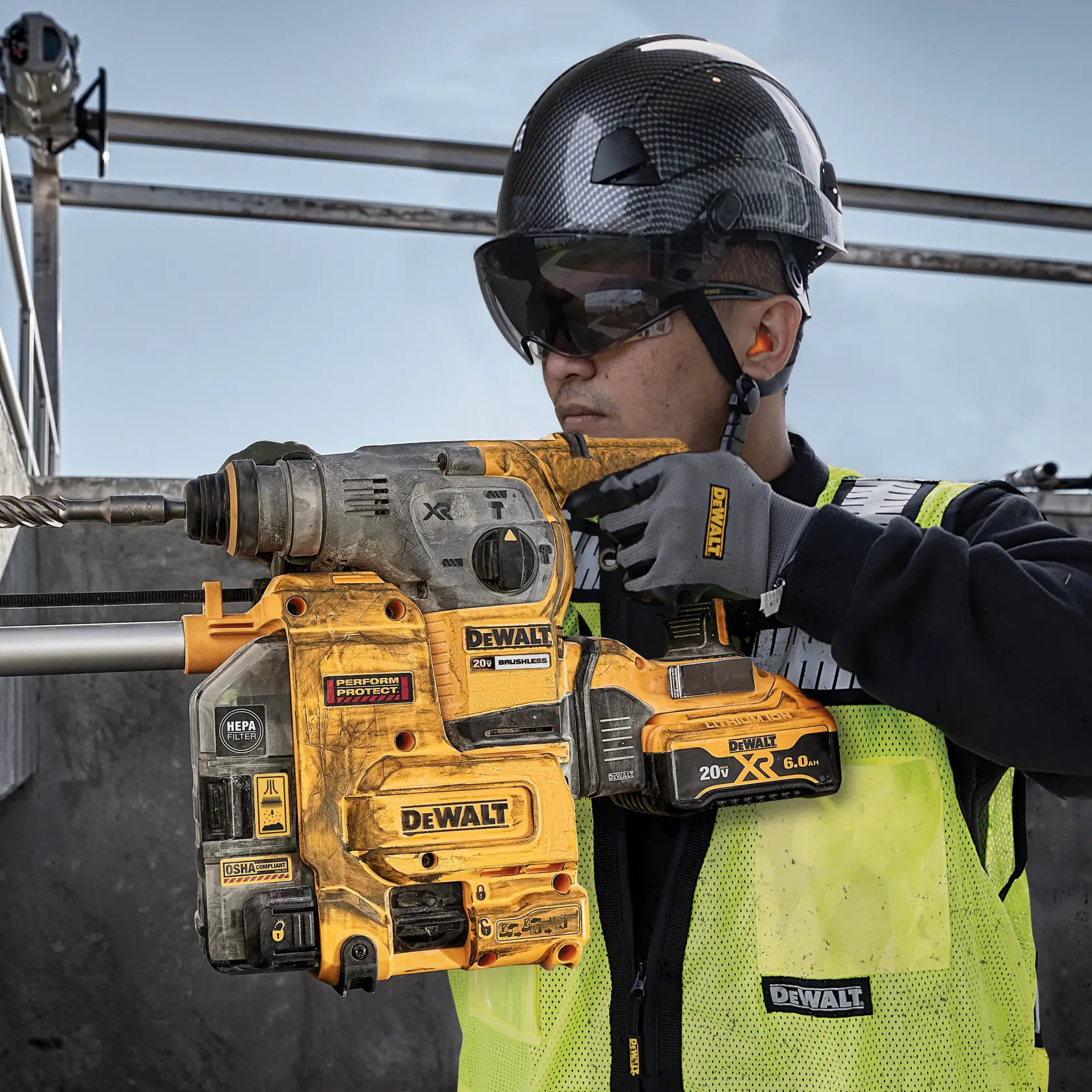 WORKER ON JOBSITE OPERATING A DEWALT POWER TOOL WEARING A SLATE COLOR TYPE II HELMET, SAFETY GLASSES UNDER A SMOKE TINTED VISOR AND ORANGE EARPLUGS