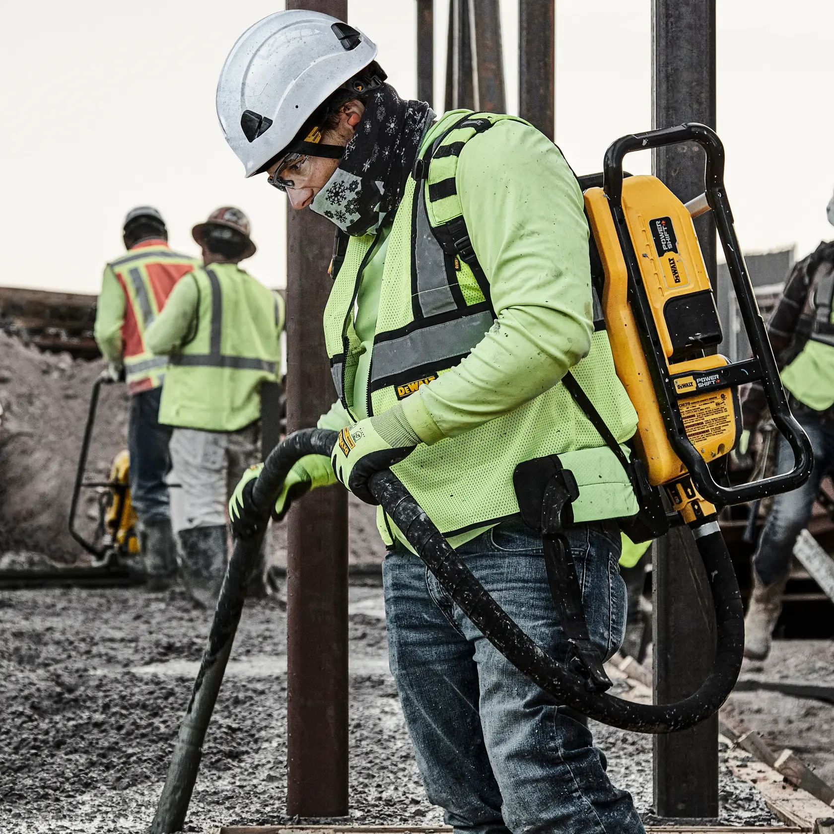 A construction worker wearing a safety helmet, high-visibility vest, and gloves is operating a DEWALT portable power tool on a muddy construction site. The worker has a DEWALT device strapped to their back. Other workers are visible in the background also wearing protective gear.