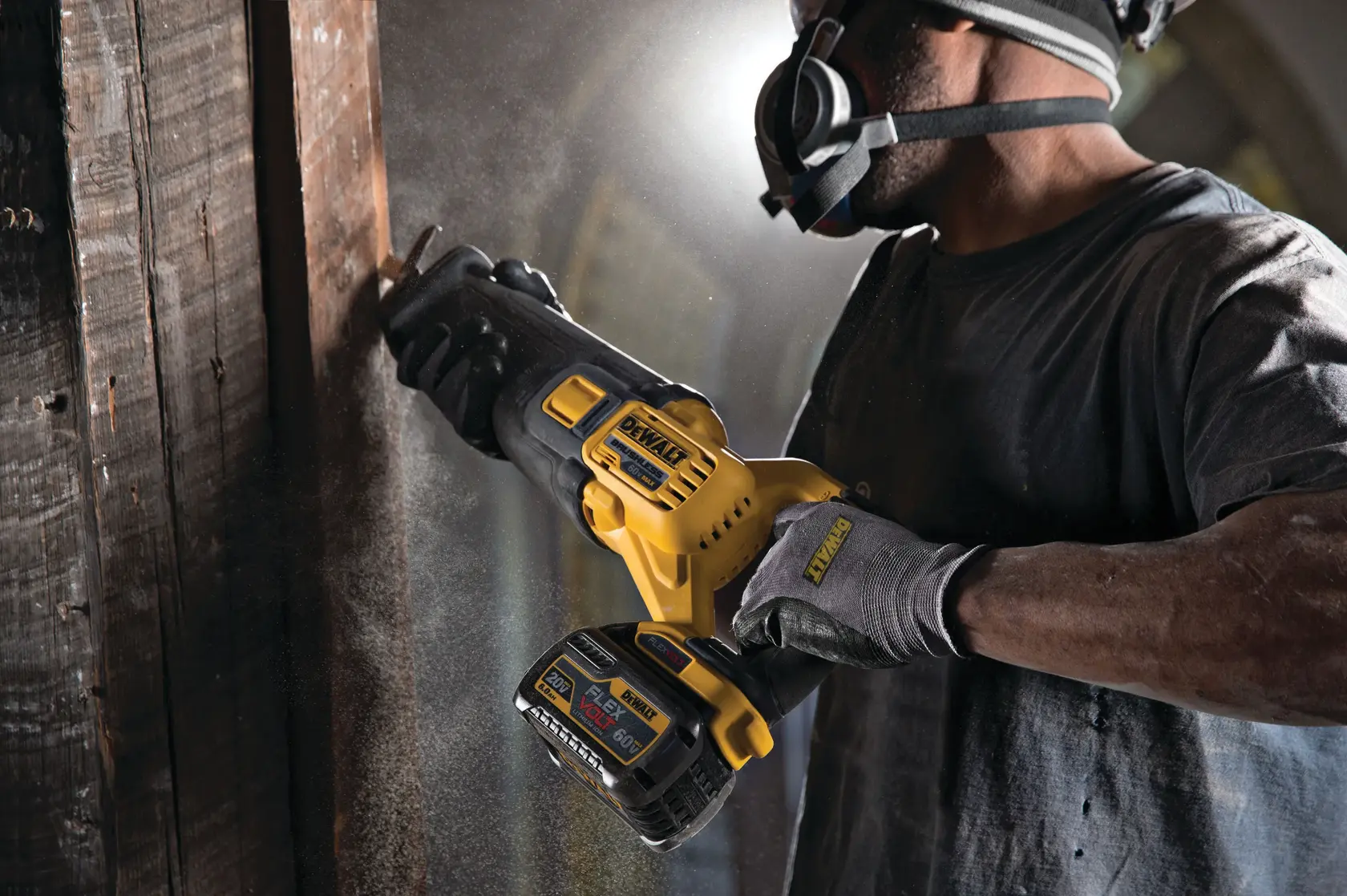 A person wearing protective gloves and a respirator mask is using a yellow and black DEWALT cordless reciprocating saw to cut through a piece of wood indoors. Sawdust is visible in the air.