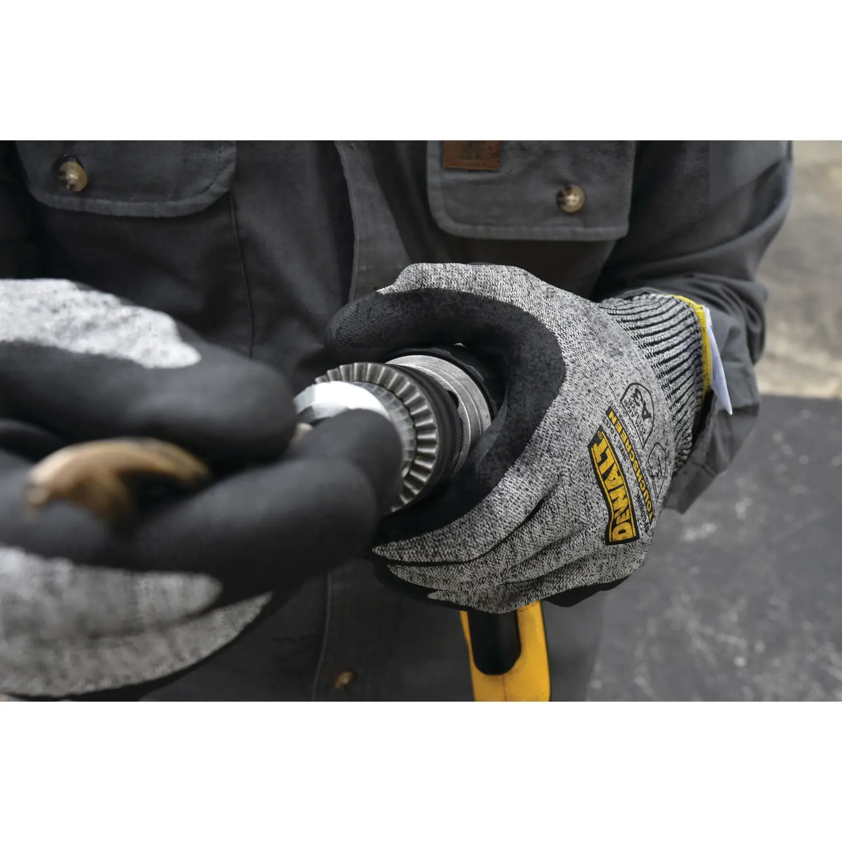 A close-up of a person wearing DEWALT protective work gloves, holding a tool or an industrial hose fitting. The gloves are gray and black with DEWALT branding, and the person is wearing a dark work jacket.