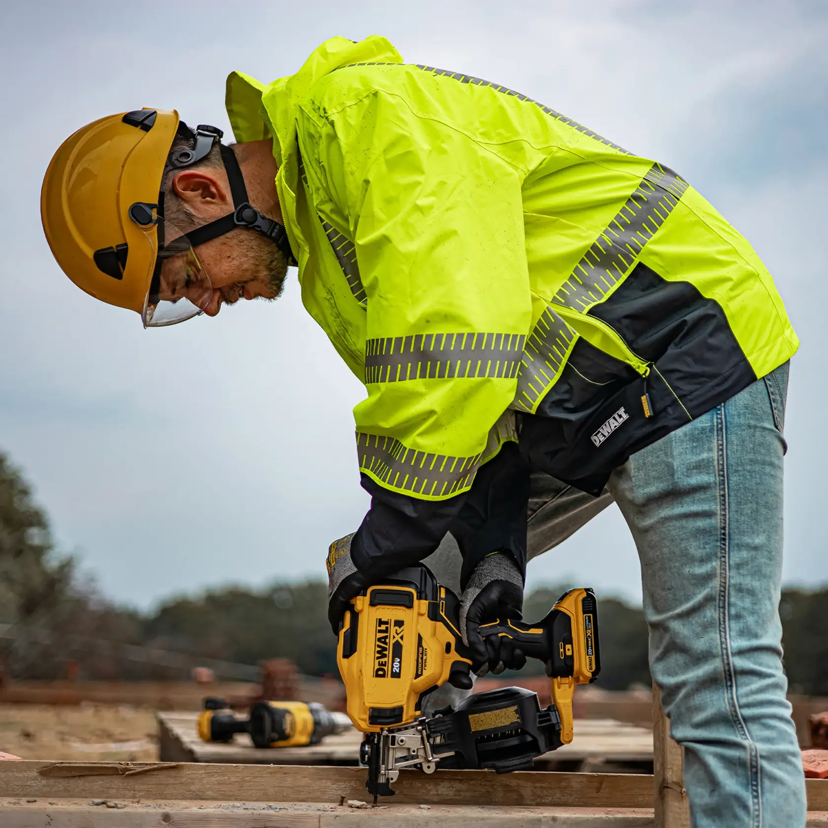 A person wearing a yellow safety jacket and helmet is using a DEWALT cordless power tool outdoors at a construction site.