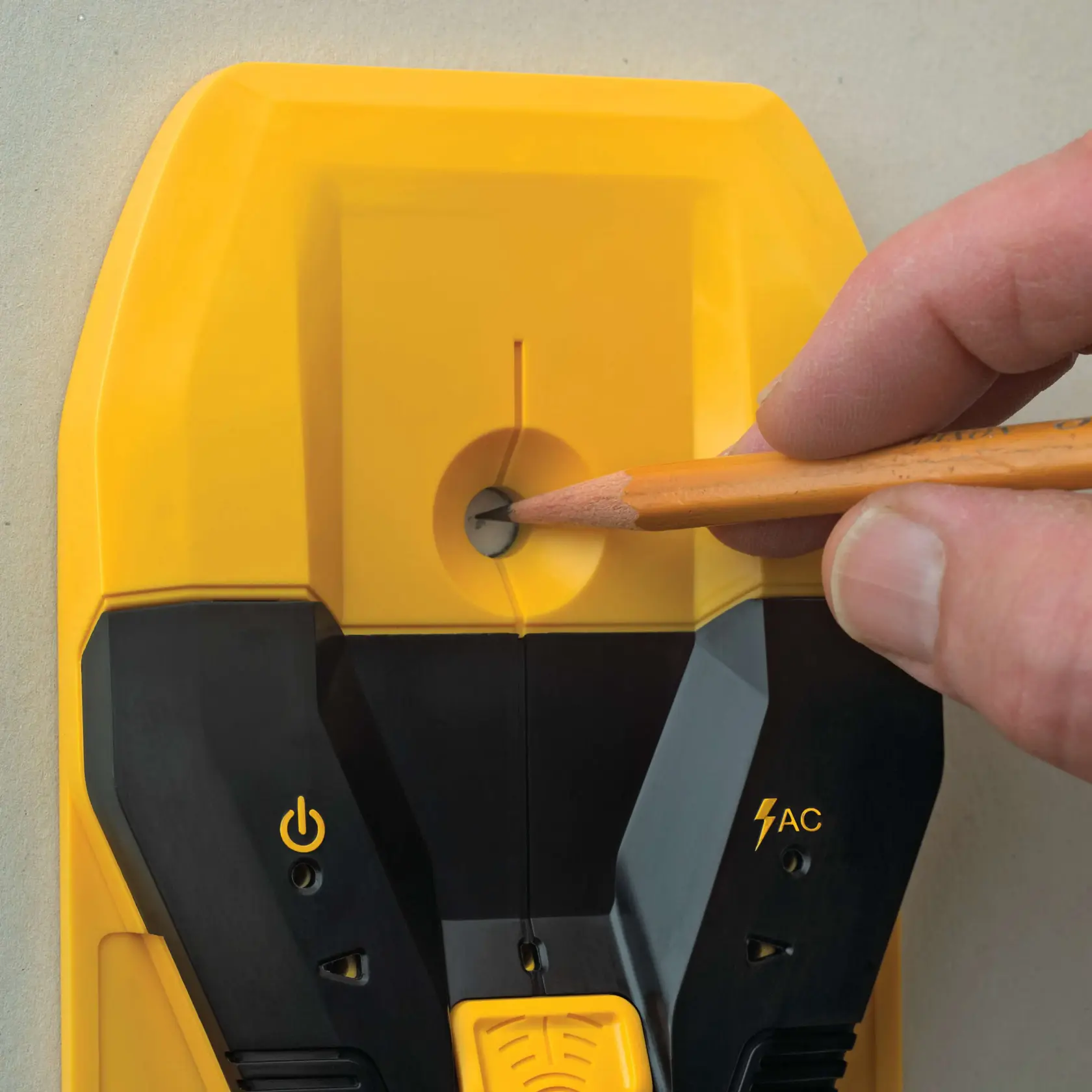 A close-up view of a DEWALT stud finder being used on a wall, with a hand holding a pencil marking the location through a round marking slot on the tool.