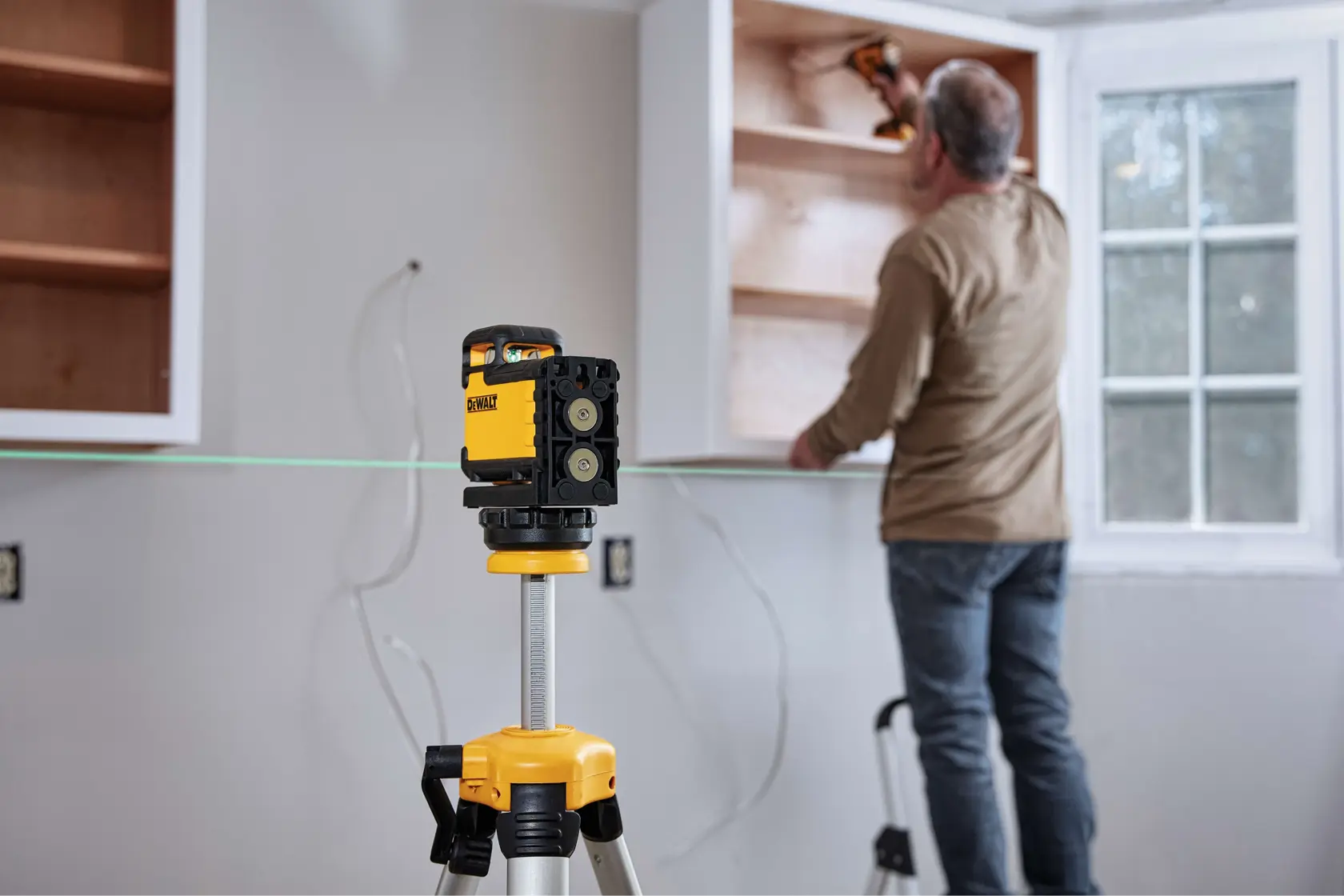A DEWALT green line laser level is set up on a tripod in a room under construction, emitting a green laser across the space. A person with a blurred face is working in the background near cabinets and wall outlets.