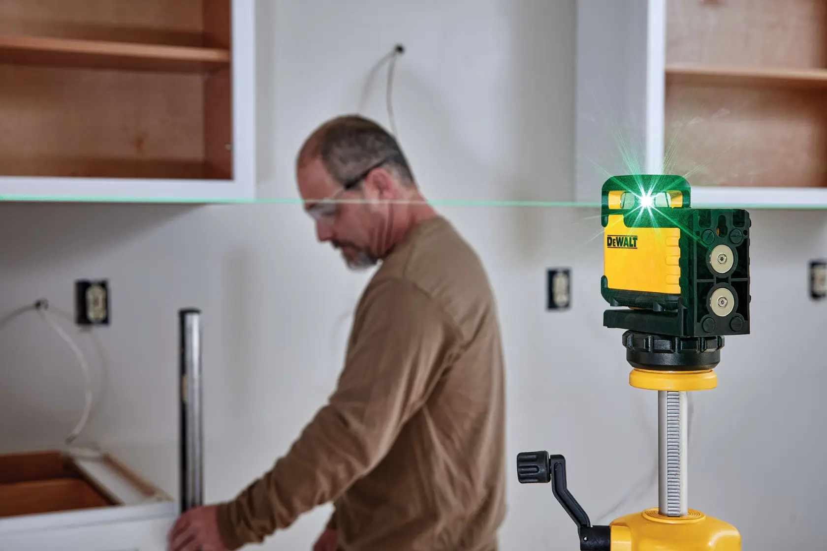 A DEWALT laser level device is mounted on a tripod in the foreground, with a person installing white cabinets in a kitchen under construction. The walls have exposed wires and there are open cabinet frames and tools visible.