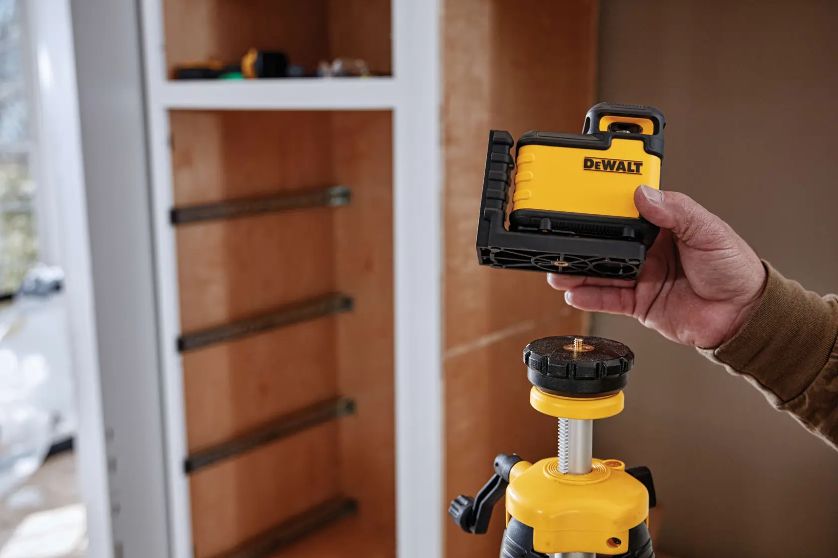 A person is holding a DEWALT laser level device, about to attach it onto a tripod. The scene is set indoors near a cabinet under construction.