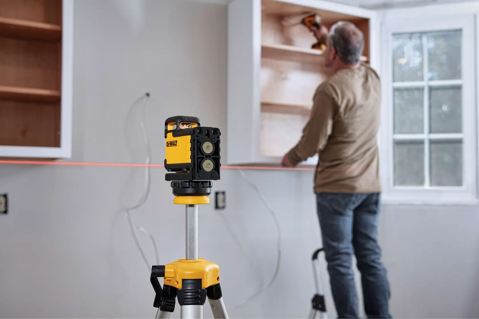 A DEWALT laser level tool mounted on a tripod is positioned in the foreground, projecting a red laser line across a wall. In the background, a person is installing or adjusting a cabinet with a power tool in a room with a window.