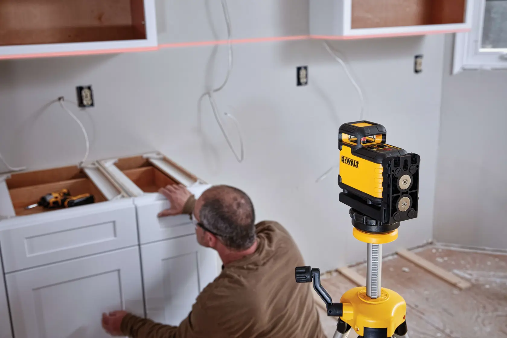 A DEWALT laser level device is set up on a tripod in a room under renovation, helping align cabinets being installed by a worker.