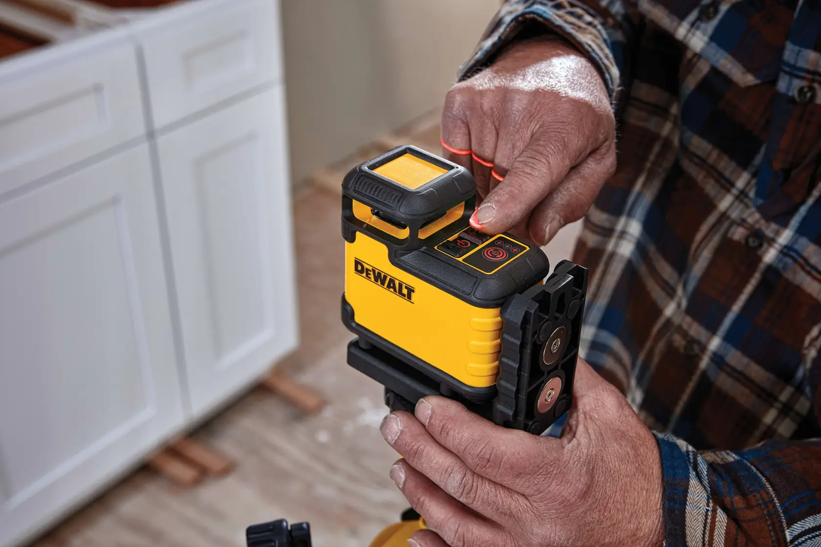 Close-up of a person operating a yellow DEWALT laser level device, which is positioned on a mounting bracket. The person's finger is pressing a button on the top panel. Nearby, a kitchen cabinet and a plaid shirt sleeve are visible.