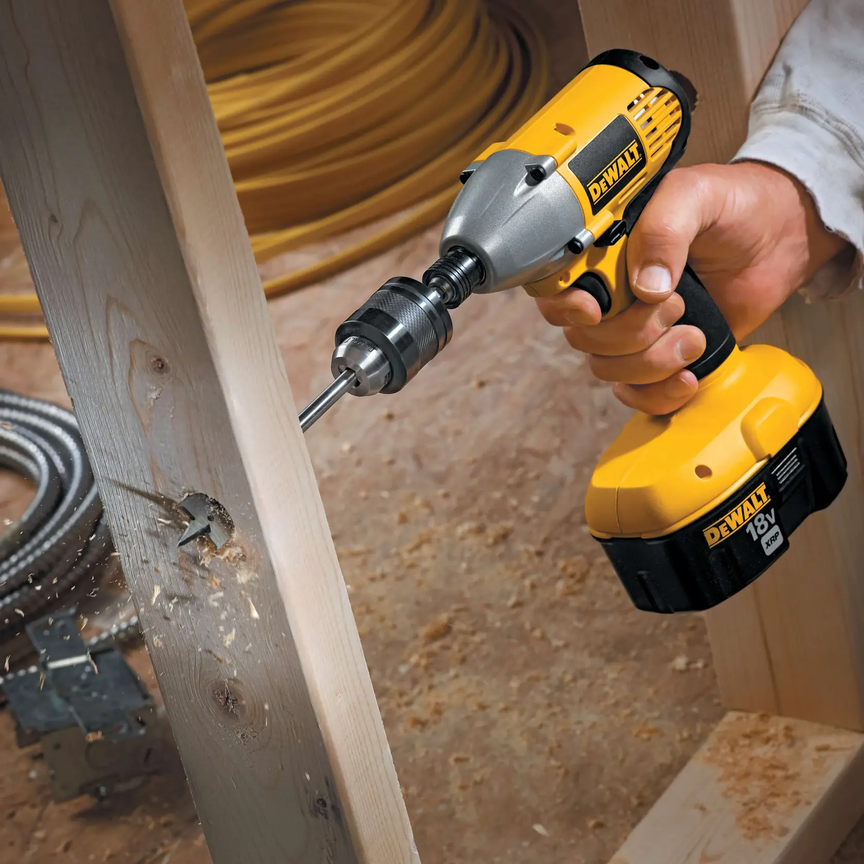 A DEWALT 18V cordless power drill is being used to drill a hole in a wooden beam. Sawdust is visible as the drill bit cuts through the wood, and there are construction materials in the background.
