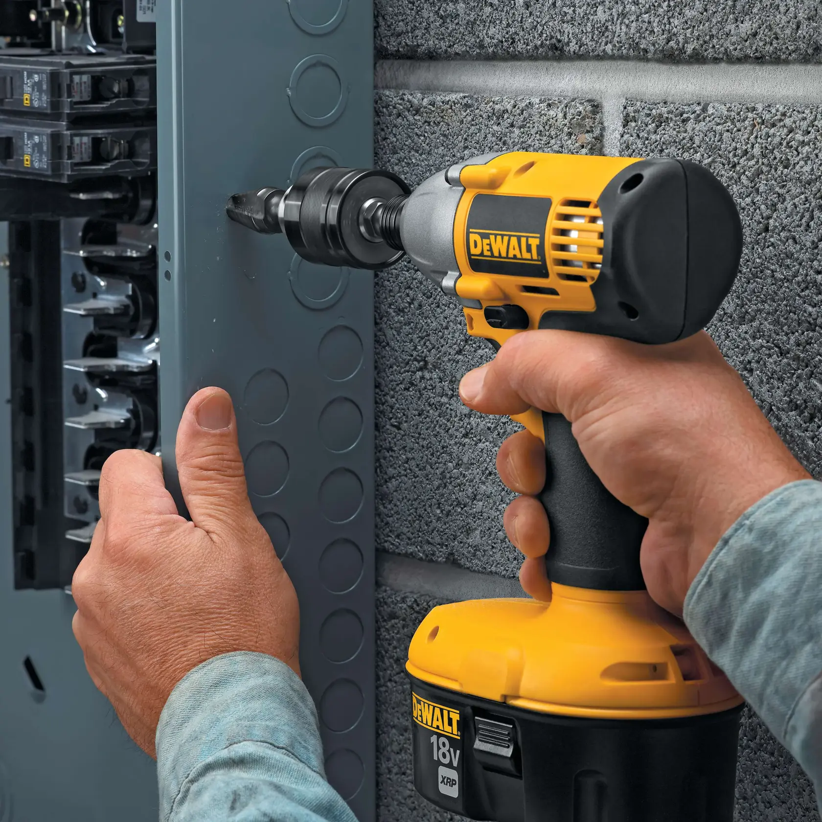 A person is using a DEWALT 18V cordless power drill with a hole saw attachment to drill into a metal electrical panel. The drill is held firmly and positioned next to the panel, which is mounted on a concrete block wall.