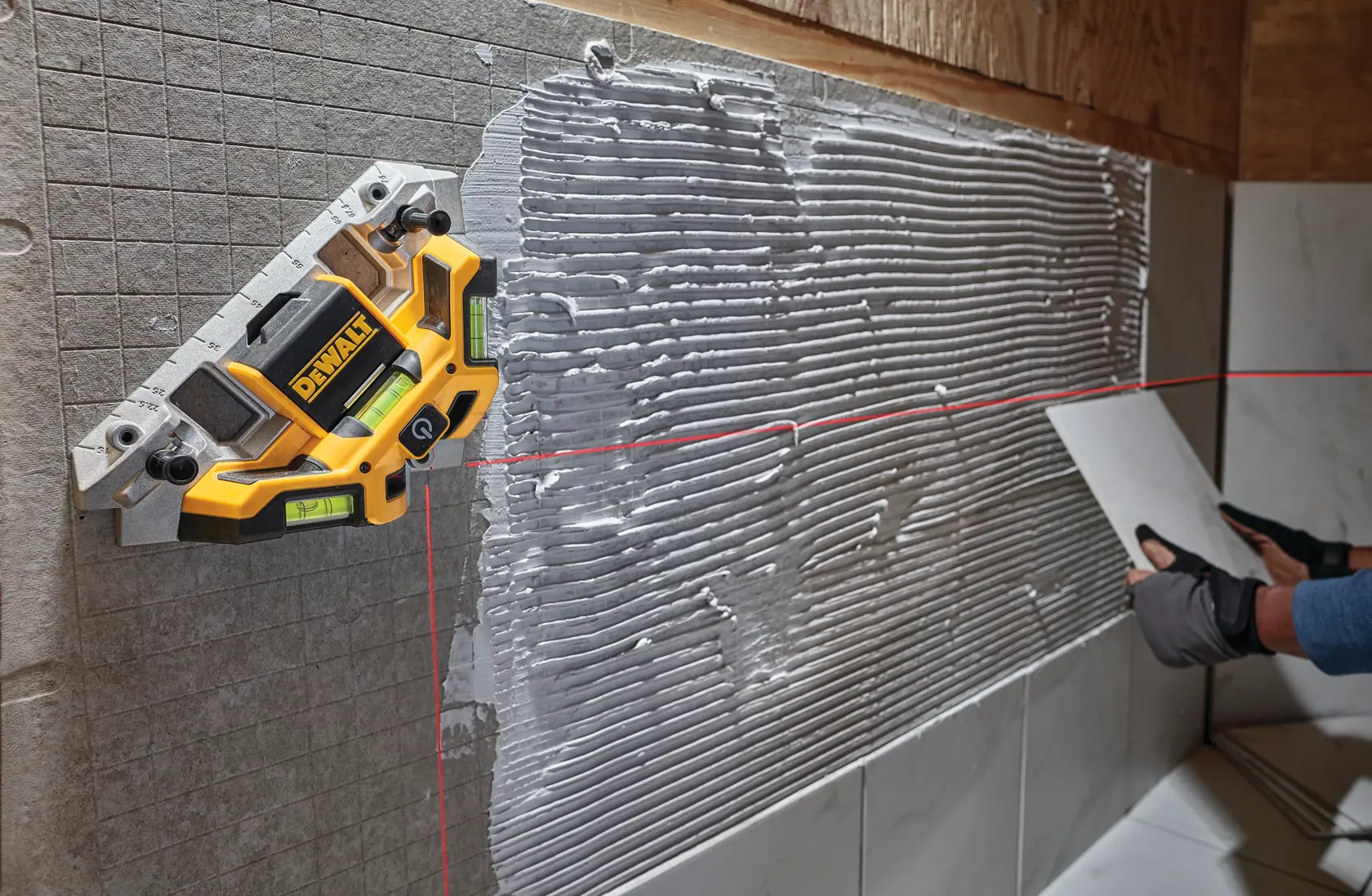 A DEWALT laser level (model DW0802) is attached to a tiled wall, projecting horizontal and vertical red laser lines to help with alignment. The wall has adhesive applied, and a gloved hand is holding a tile, preparing for installation.