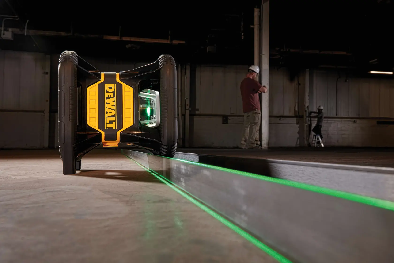 A DEWALT green line laser level is positioned on the floor in an industrial setting, projecting a bright green laser line along a metal surface. Two people in safety gear are visible in the background.
