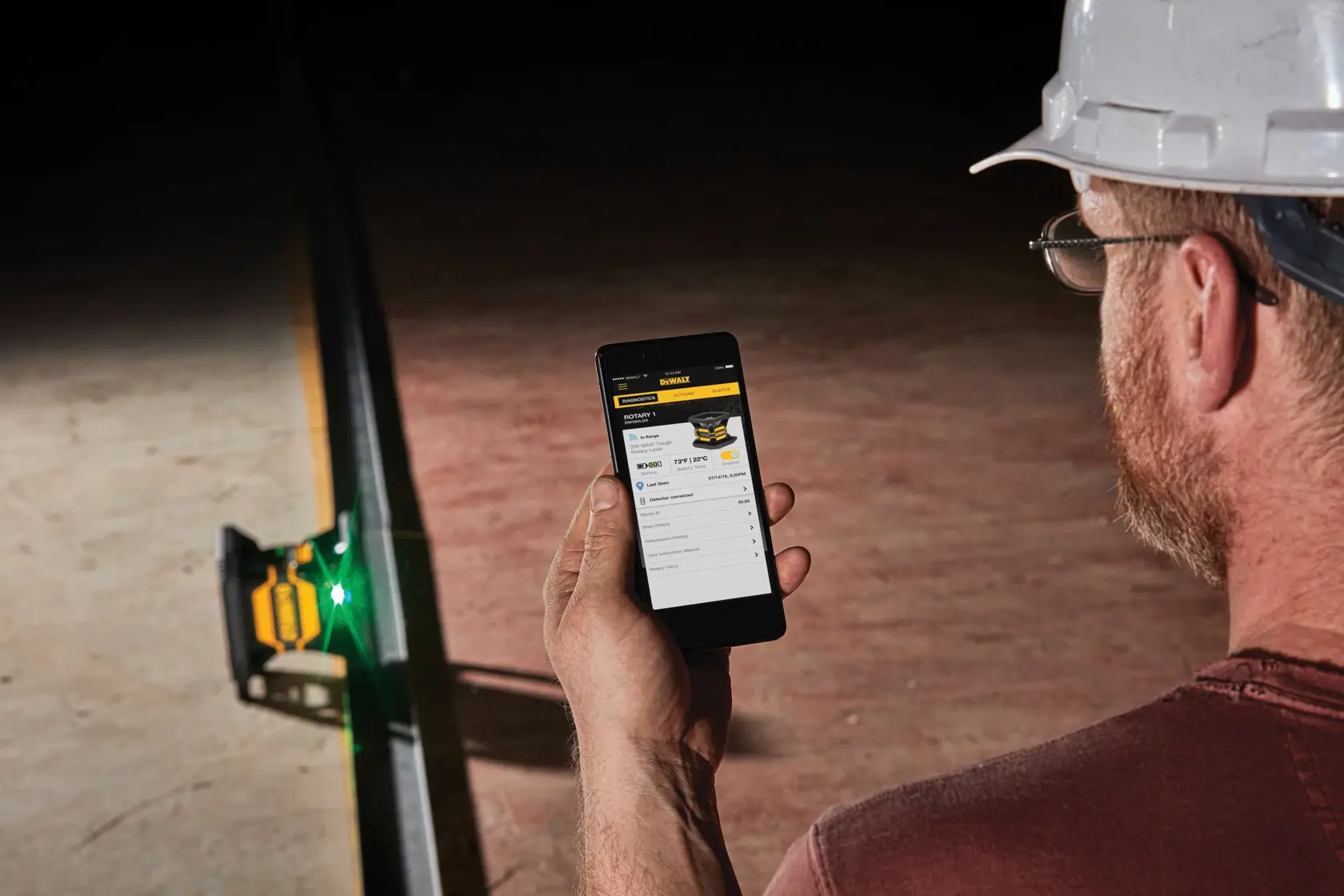 A person wearing a white hard hat is holding a smartphone and using it to control or monitor a DEWALT laser device placed on the ground, which emits a visible green laser line. The setting appears to be a construction or industrial environment.