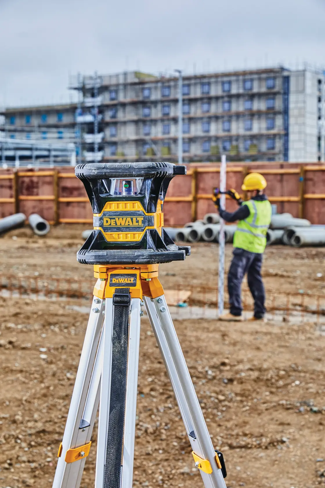 A DEWALT DW080LGS laser level mounted on a tripod at a construction site, with a worker in a high-visibility vest and yellow helmet measuring in the background.