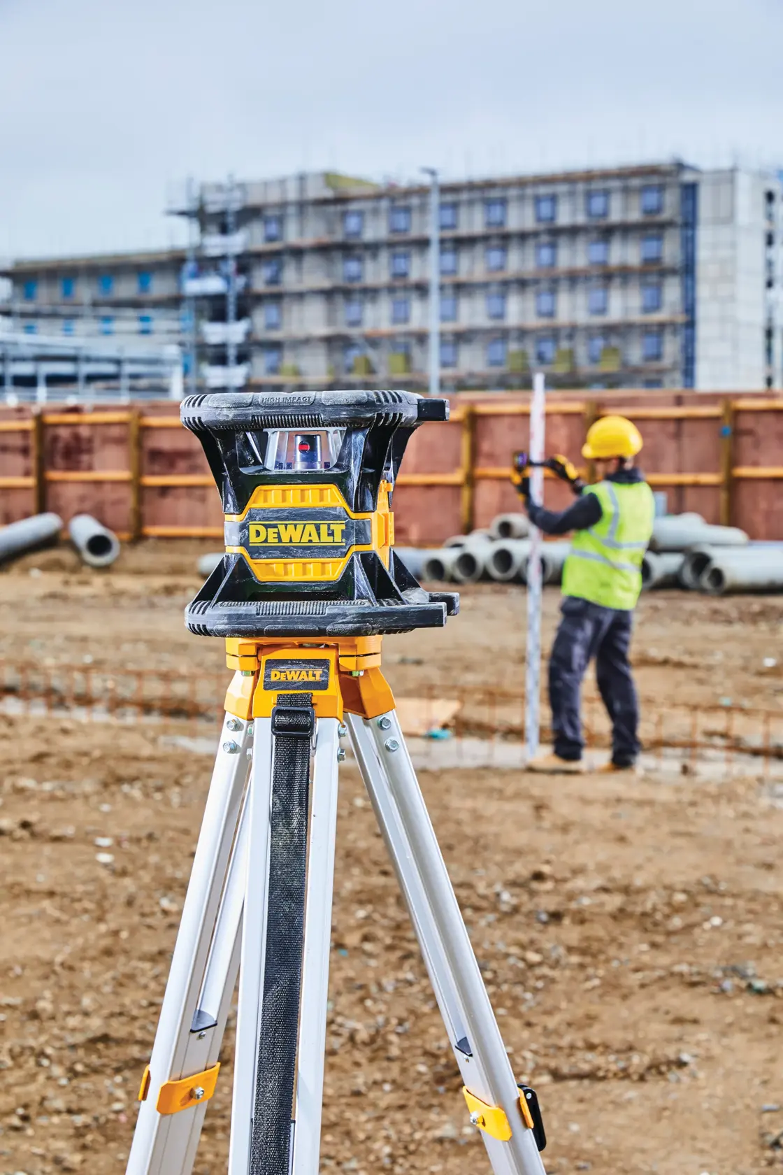 A DEWALT laser level mounted on a tripod at a construction site, with a worker in a safety vest and helmet using a measuring rod in the background. Pipes and a building under construction are visible.