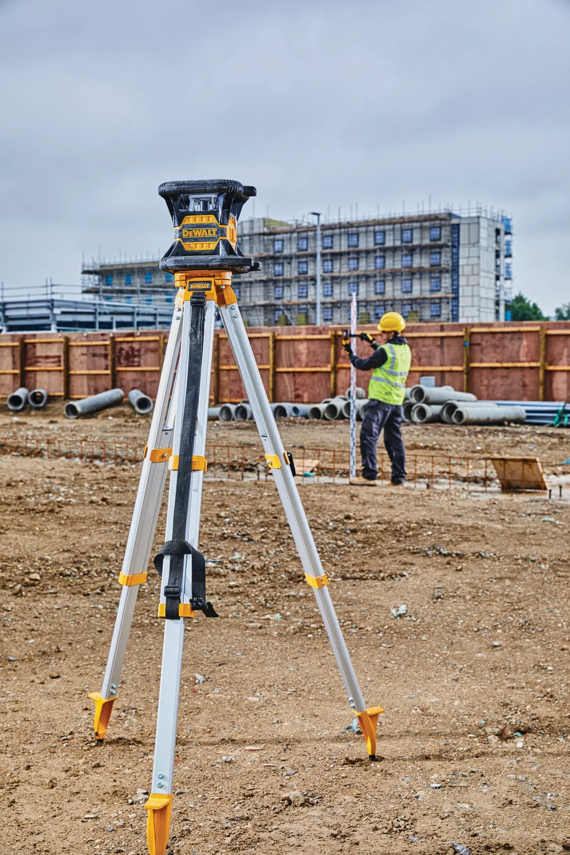 A DEWALT rotary laser level mounted on a tripod at a construction site. A worker in a yellow safety vest and helmet stands in the background, and a gloved hand holds a smartphone showing the DEWALT interface in the foreground.