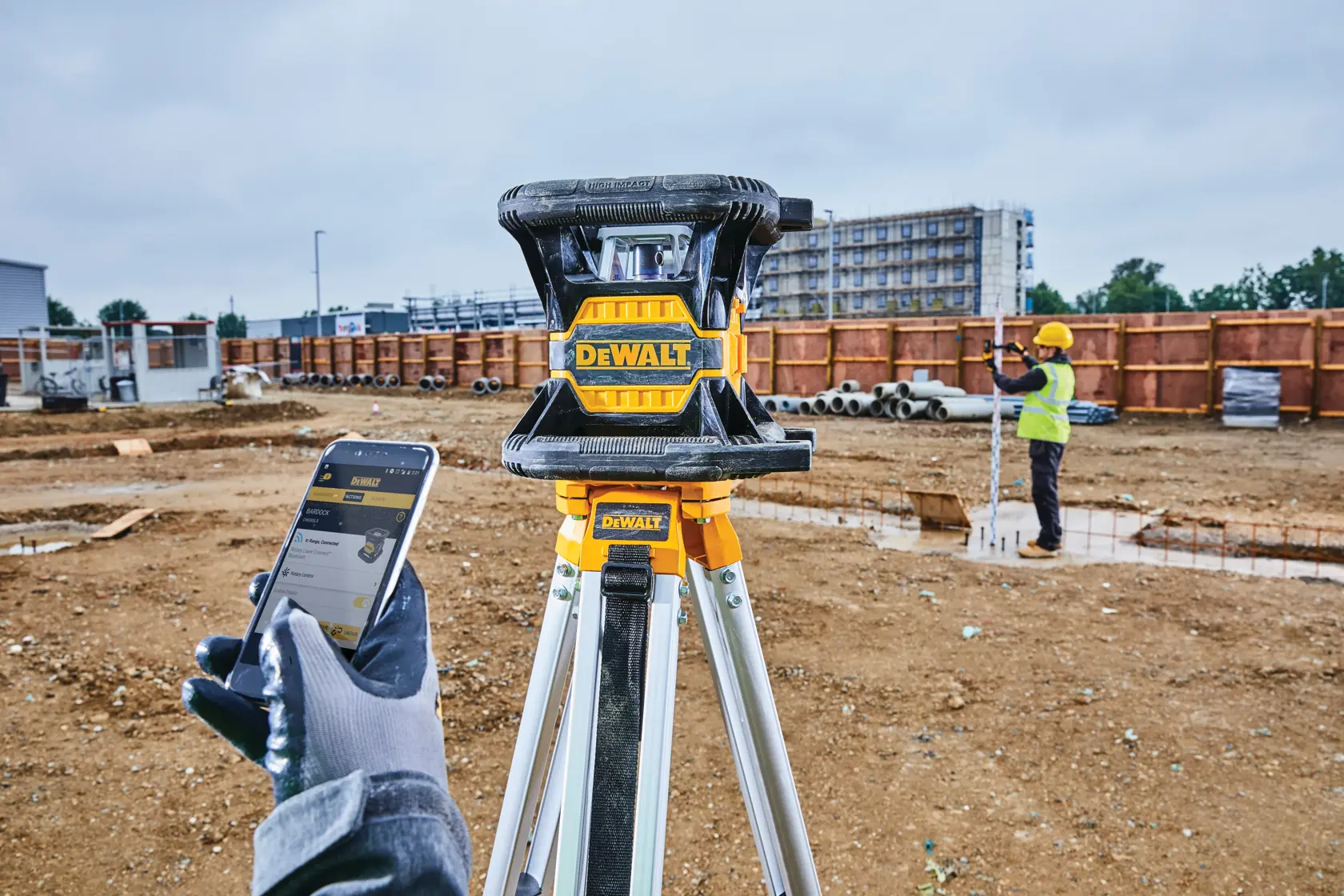 A DEWALT laser level device mounted on a tripod is shown in the foreground at a construction site. In the background, a worker wearing a safety vest and hard hat is using measuring equipment, with a building under construction visible.