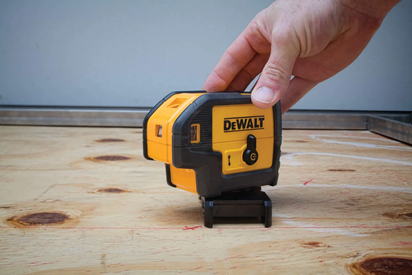 A person’s hand is adjusting a DEWALT DW085K line laser level on a wooden surface. The laser level is yellow and black and is used for precise alignment in construction or photography setups.