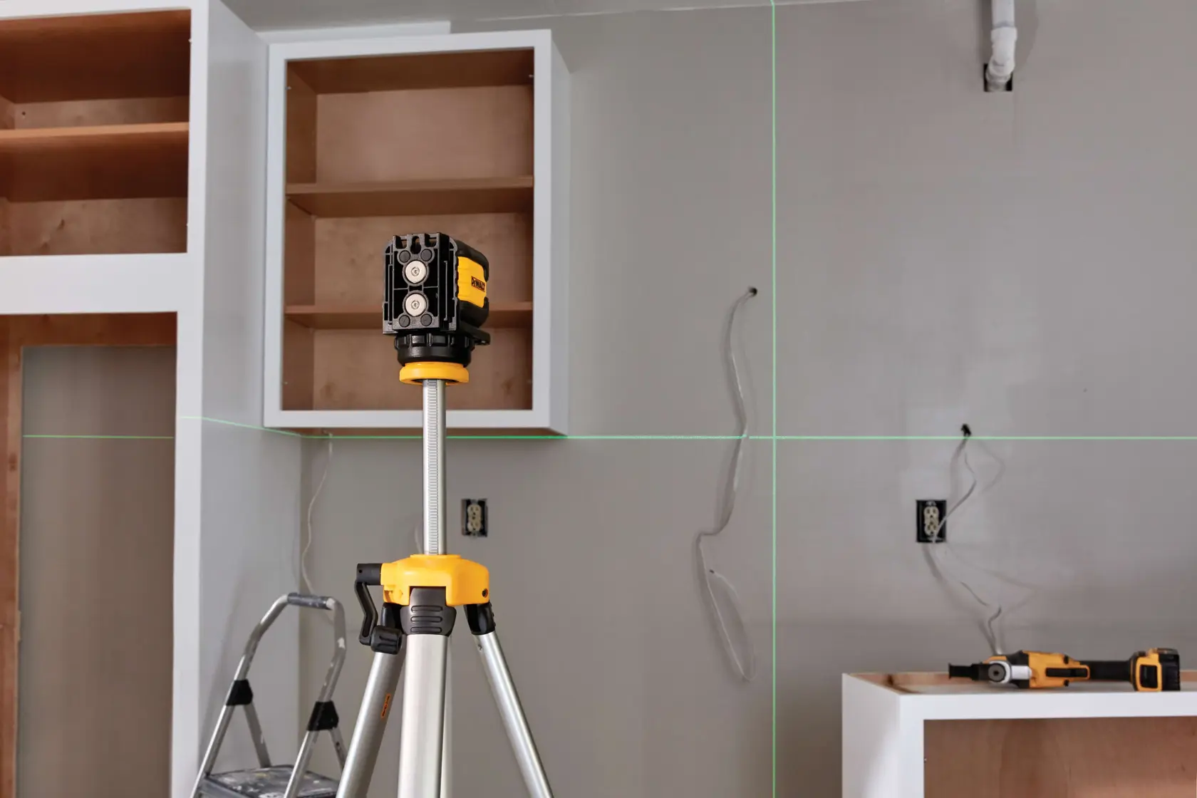 A DEWALT laser level on a tripod is positioned in a room under construction, projecting green horizontal and vertical laser lines onto the walls. The scene includes partially installed cabinets, a stepladder, wires, and DEWALT power tools resting on a nearby surface.