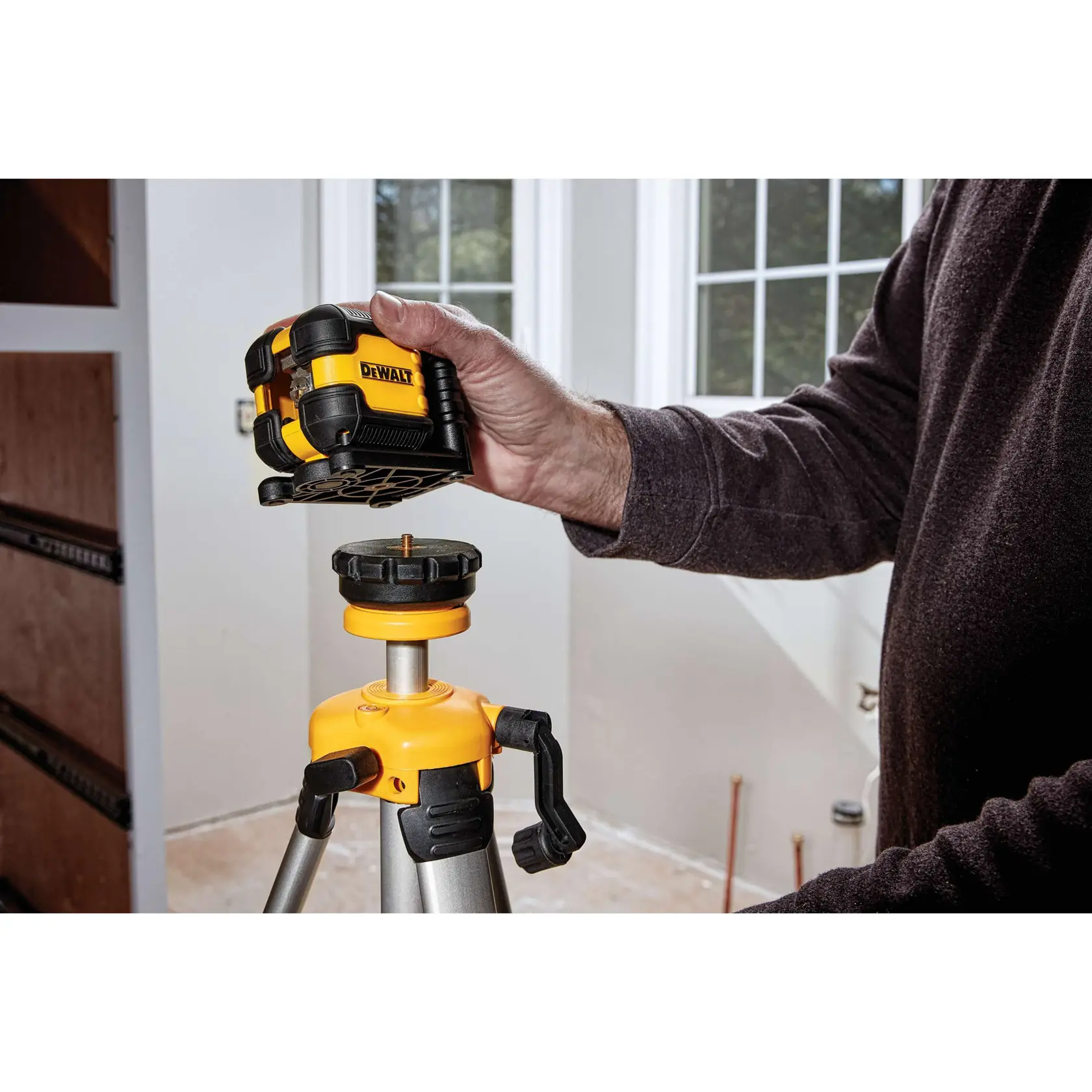 A person is placing a DEWALT DW08802 laser level onto a tripod inside a room with windows.