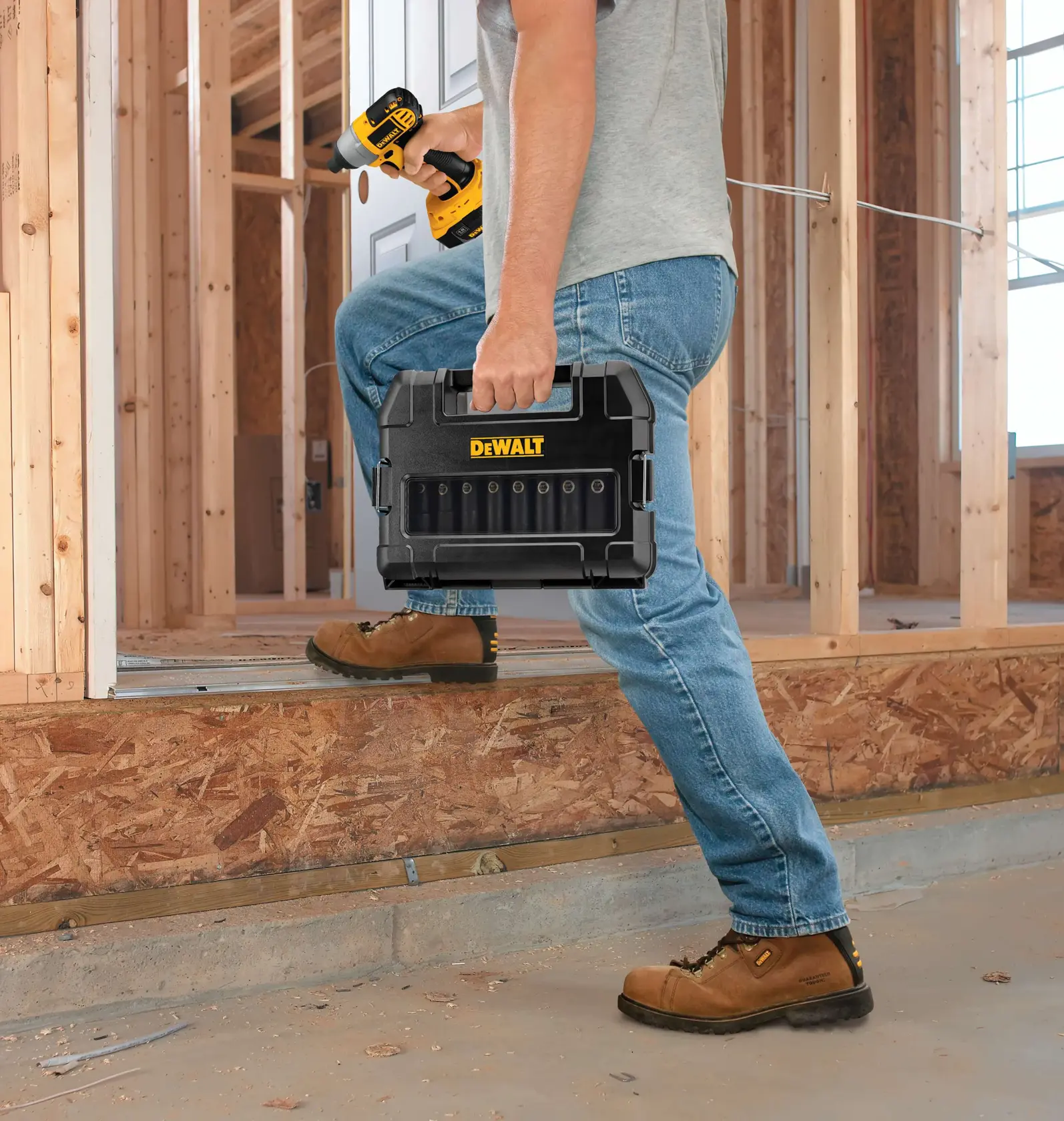 A person wearing work boots and jeans is holding a DEWALT black tool case and a yellow power drill while stepping into a construction site with exposed wooden framing.