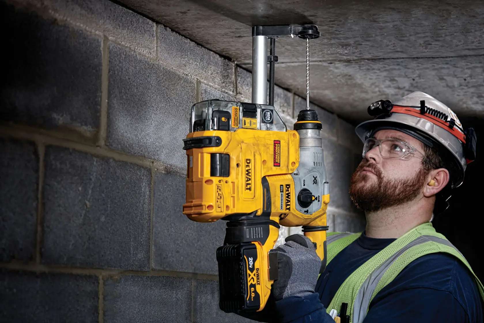 A person wearing gloves and a safety helmet uses a yellow and black DEWALT cordless rotary hammer drill, model DW5416, to drill into a concrete ceiling in an industrial setting.