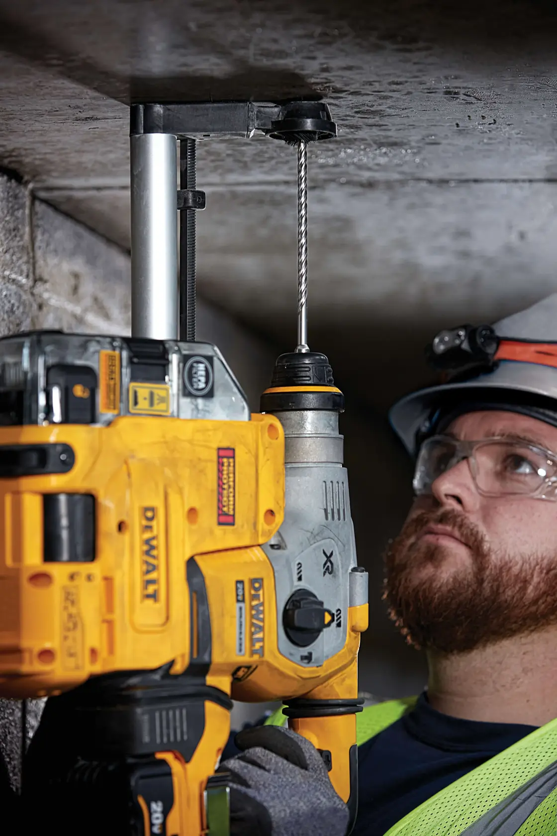 A person using a yellow DEWALT rotary hammer drill to drill into a concrete ceiling, with a dust extraction attachment connected to the tool. The individual’s face is blurred for privacy and they are wearing a safety vest and helmet.