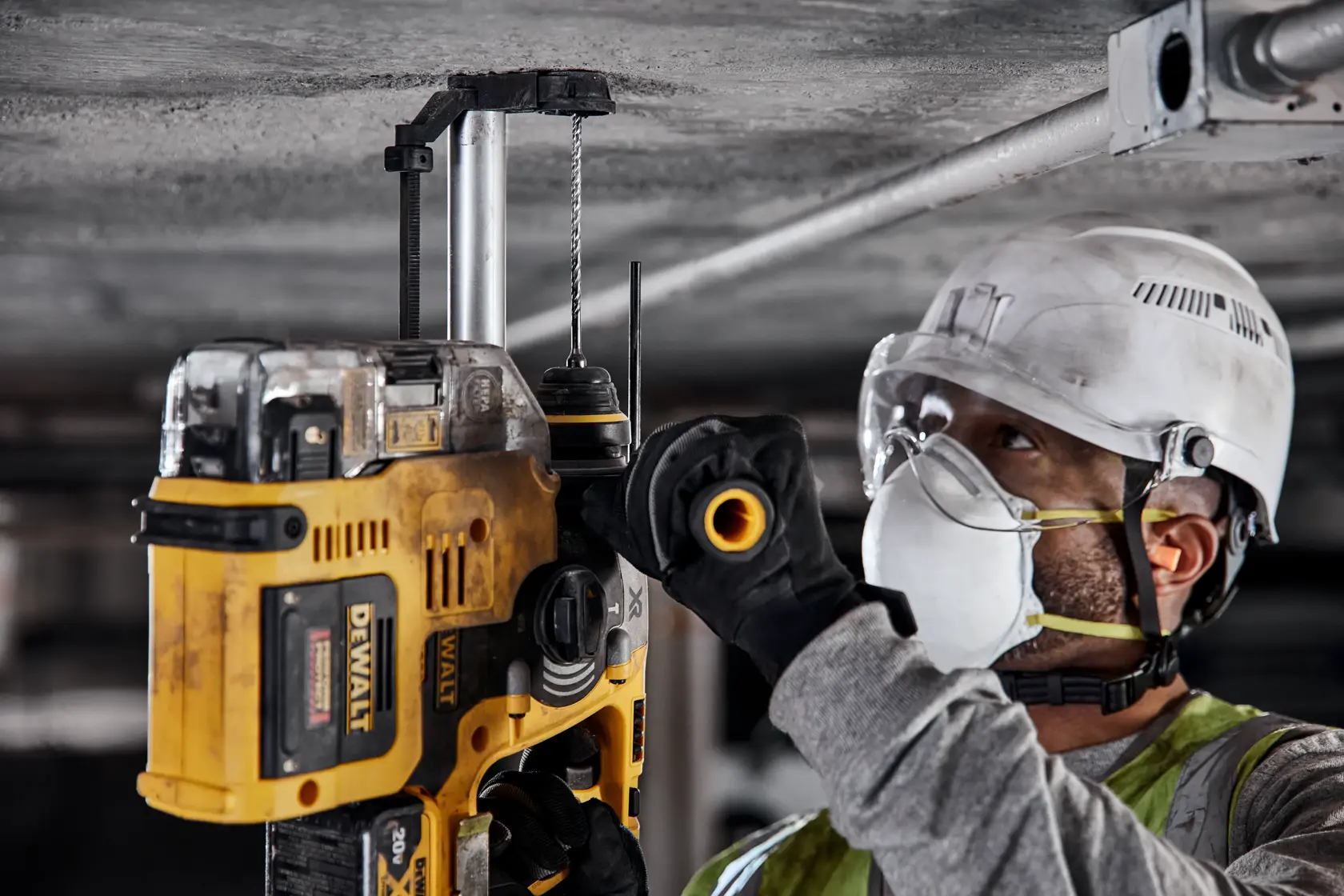 A person wearing protective gear, including a hard hat, safety goggles, gloves, and a face mask, uses a DEWALT power tool to drill into a concrete ceiling.