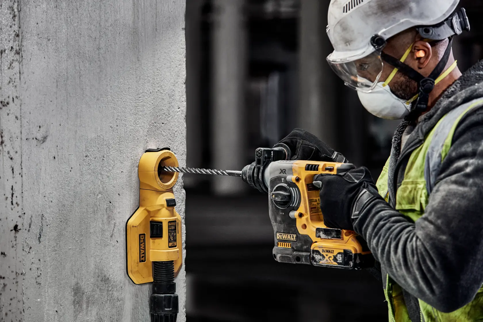 A construction worker wearing safety equipment operates a DEWALT rotary hammer drill and dust extractor against a concrete wall. The DEWALT tools are yellow and black, and the worker is focused on drilling into the wall.