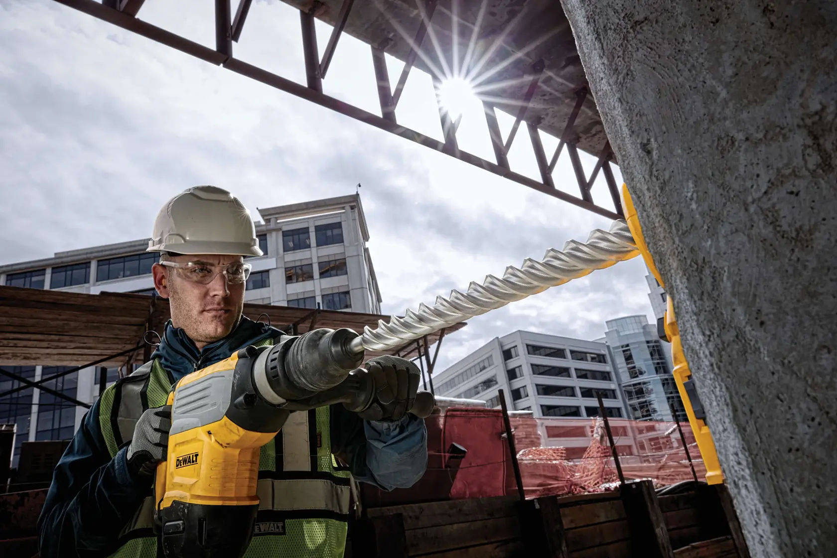 A construction worker using a DEWALT hammer drill with a large spiral drill bit to drill into concrete at an outdoor construction site. The worker is wearing a safety vest and helmet, and modern buildings are visible in the background.
