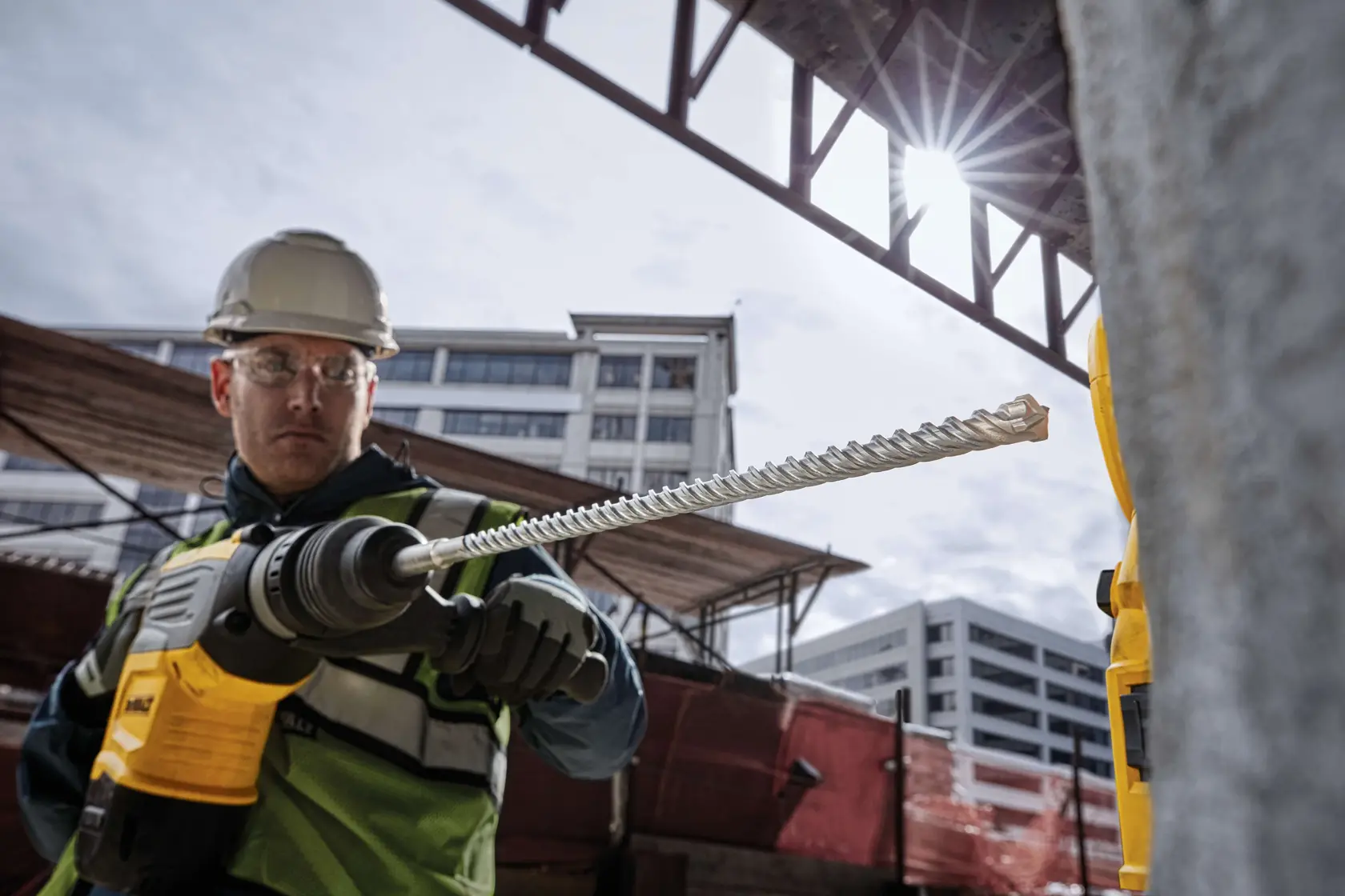 A construction worker holding a DEWALT rotary hammer drill with a long masonry drill bit, working at an outdoor construction site. The worker is wearing a safety helmet and vest, and buildings are visible in the background.