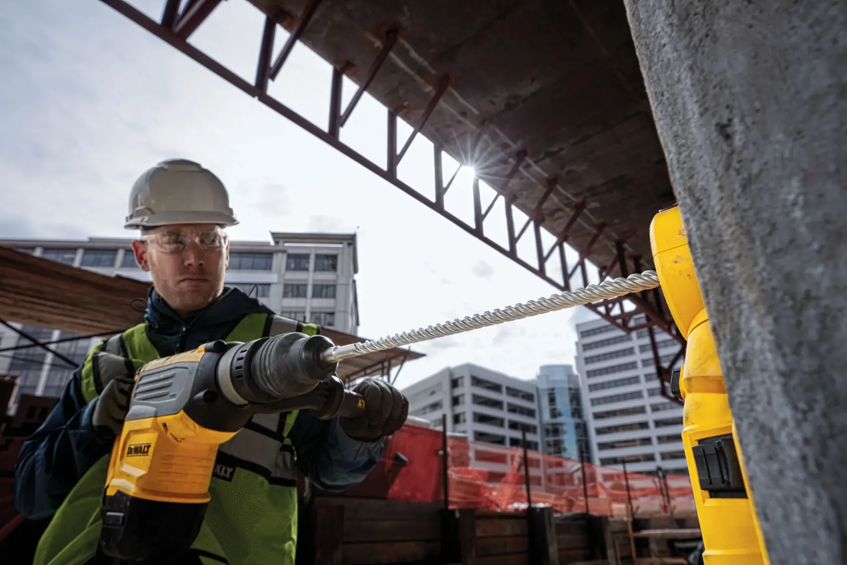 A construction worker wearing a safety vest and helmet uses a DEWALT power drill with the DW5819 drill bit to drill into concrete at an outdoor construction site. The worker's face is blurred for privacy.