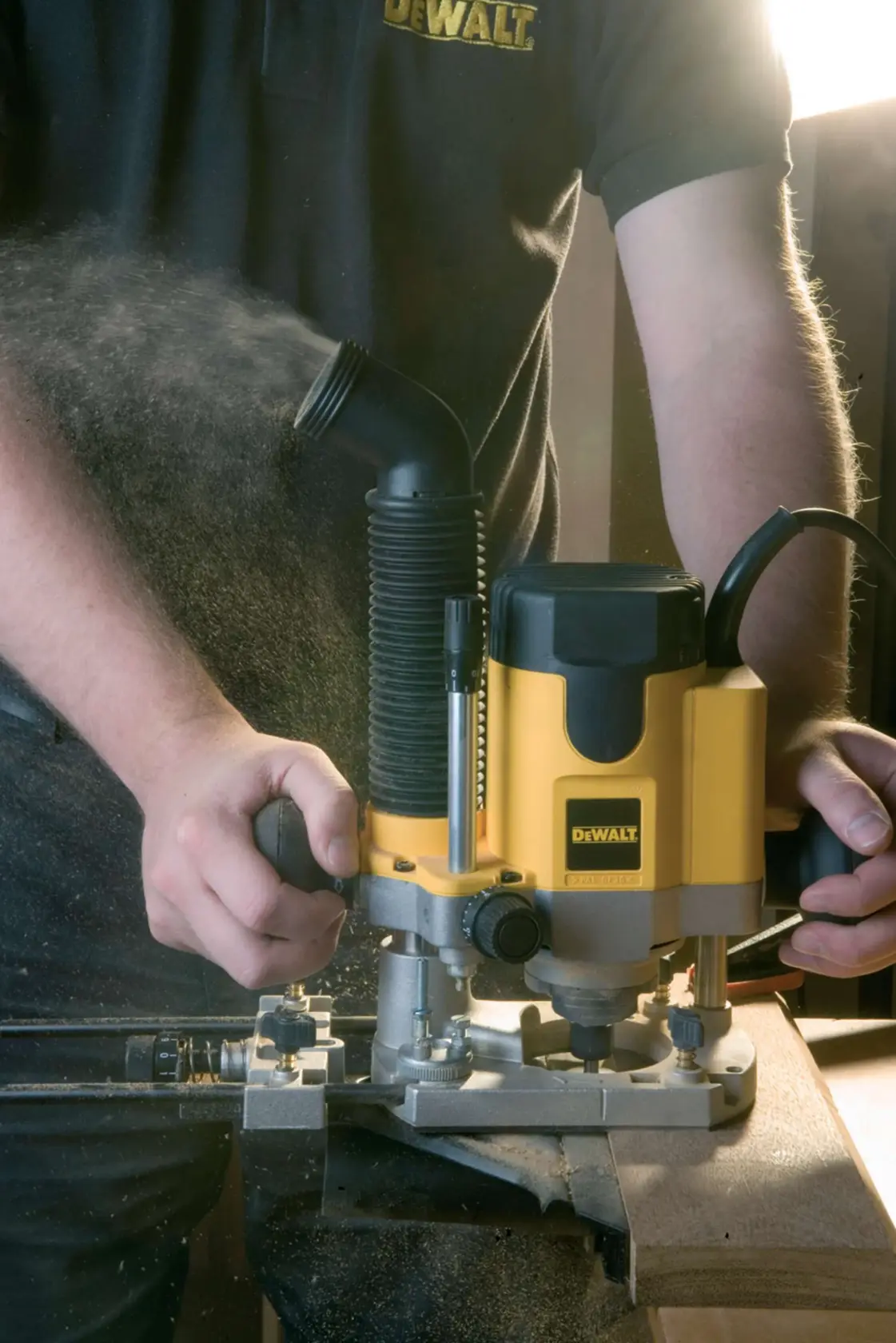 A person using a DEWALT DW621 router tool to work on a piece of wood, with wood dust being expelled through a dust extraction port. The router is yellow and black, and the operator is wearing a DEWALT-branded shirt.