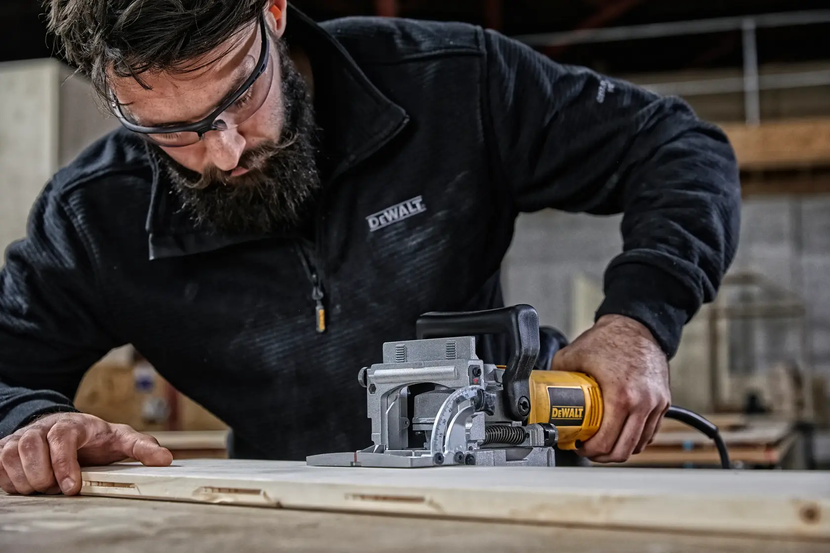 A person wearing a black DEWALT jacket uses a yellow DEWALT electric tool to cut wood in a workshop setting. The tool is positioned against a flat wooden surface, and the workspace has an industrial appearance.