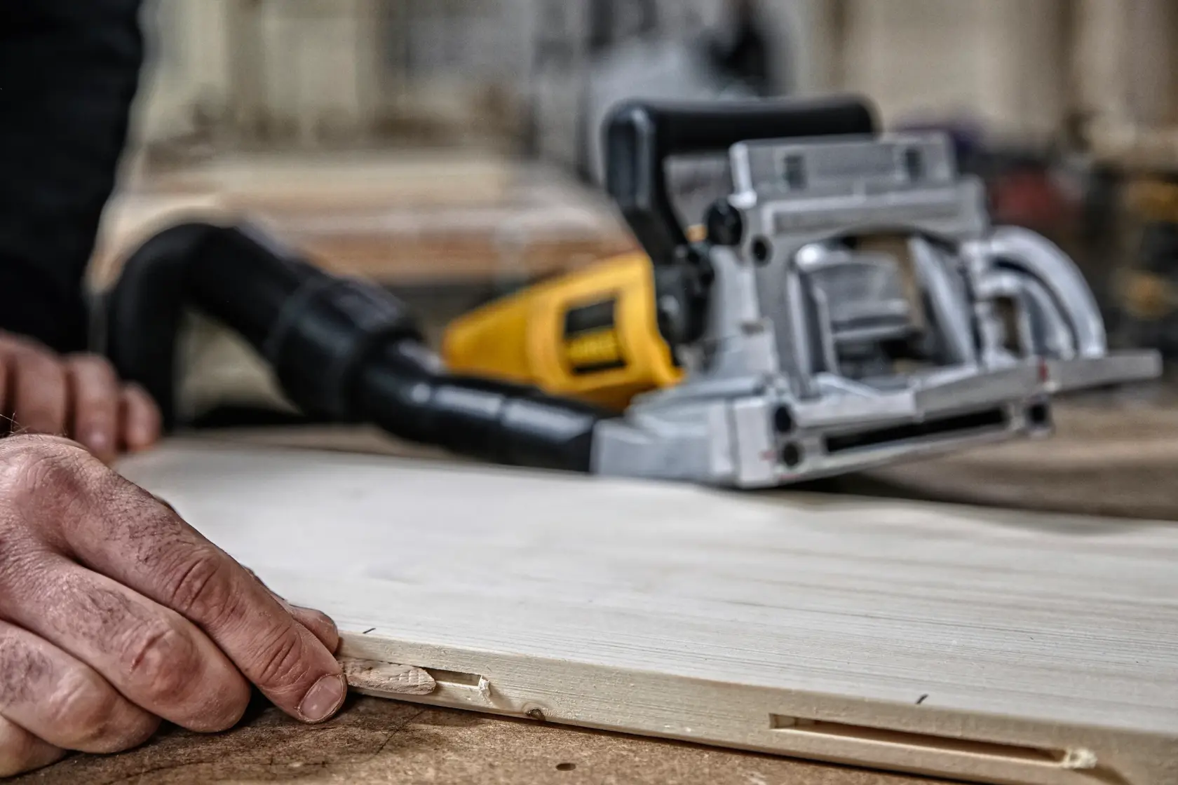 A close-up of a biscuit joiner woodworking tool and a hand placing a biscuit into a slot on a piece of wood, with a workshop setting in the background.