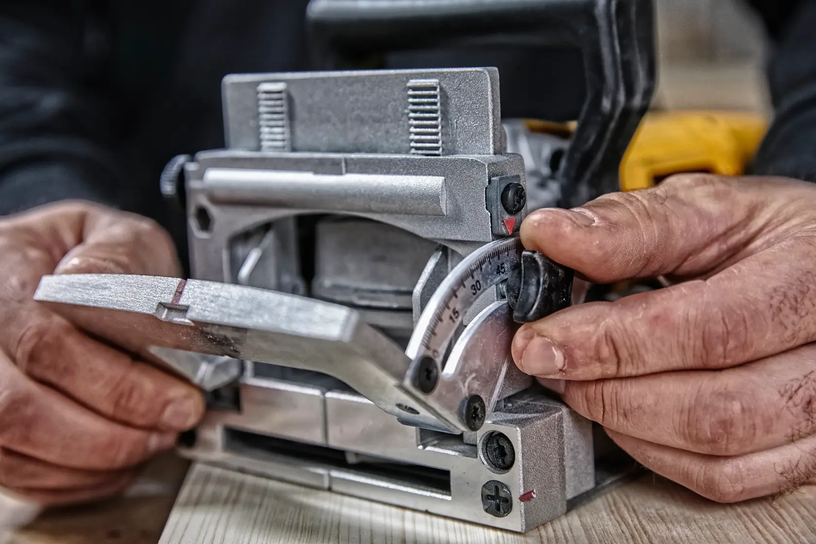 Close-up of hands adjusting the angle and settings on a biscuit joiner woodworking tool, placed on a wooden surface.