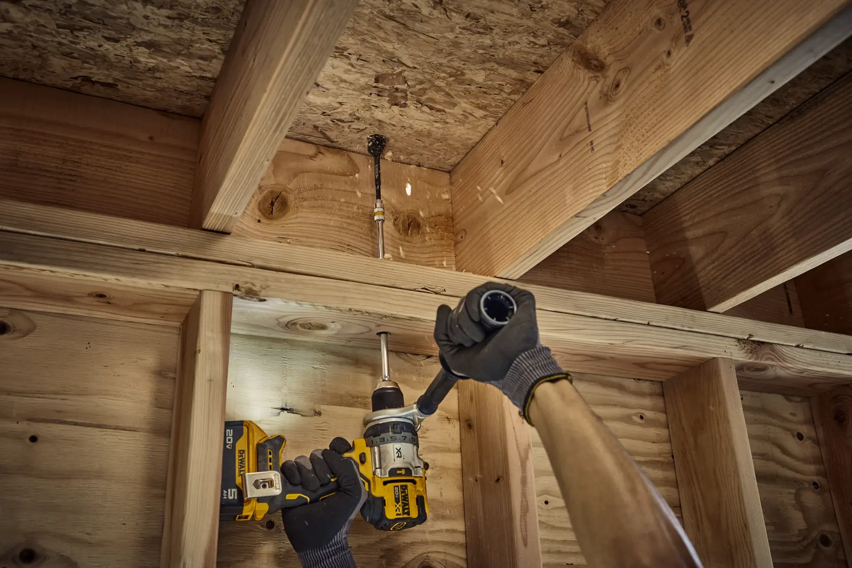 A person wearing gloves uses a DEWALT cordless drill to drill into a wooden ceiling or joist in a construction setting.