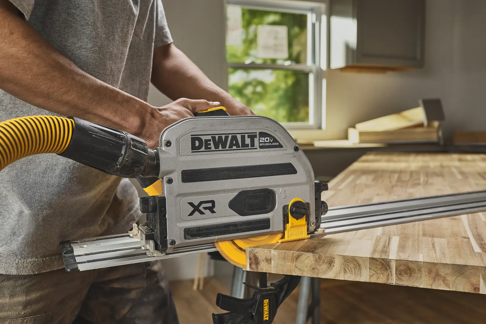A person using a DEWALT XR 20V cordless track saw with an attached dust extraction hose to cut a piece of wood on a workbench in a workshop.