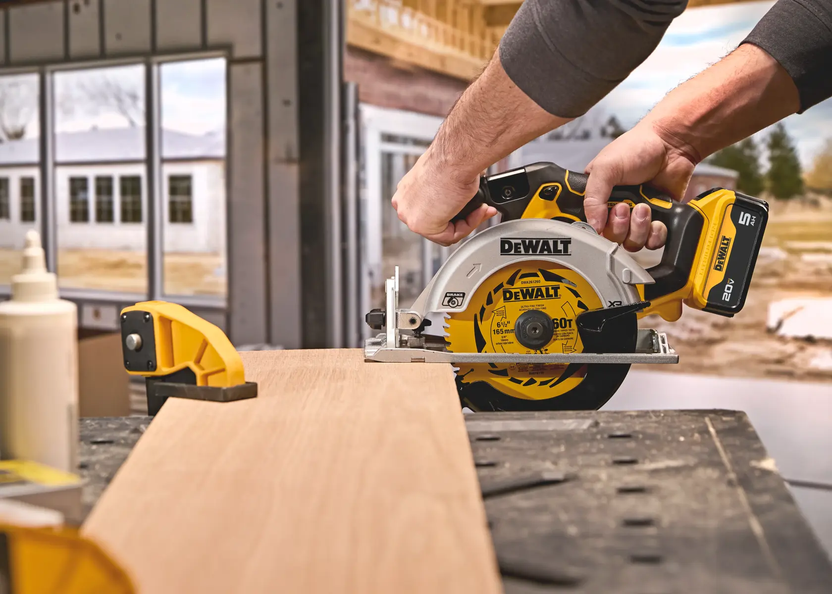A person uses a DEWALT cordless circular saw to cut a piece of wood on a workbench in a construction environment. The saw is yellow and black, and the wood is secured with a clamp.