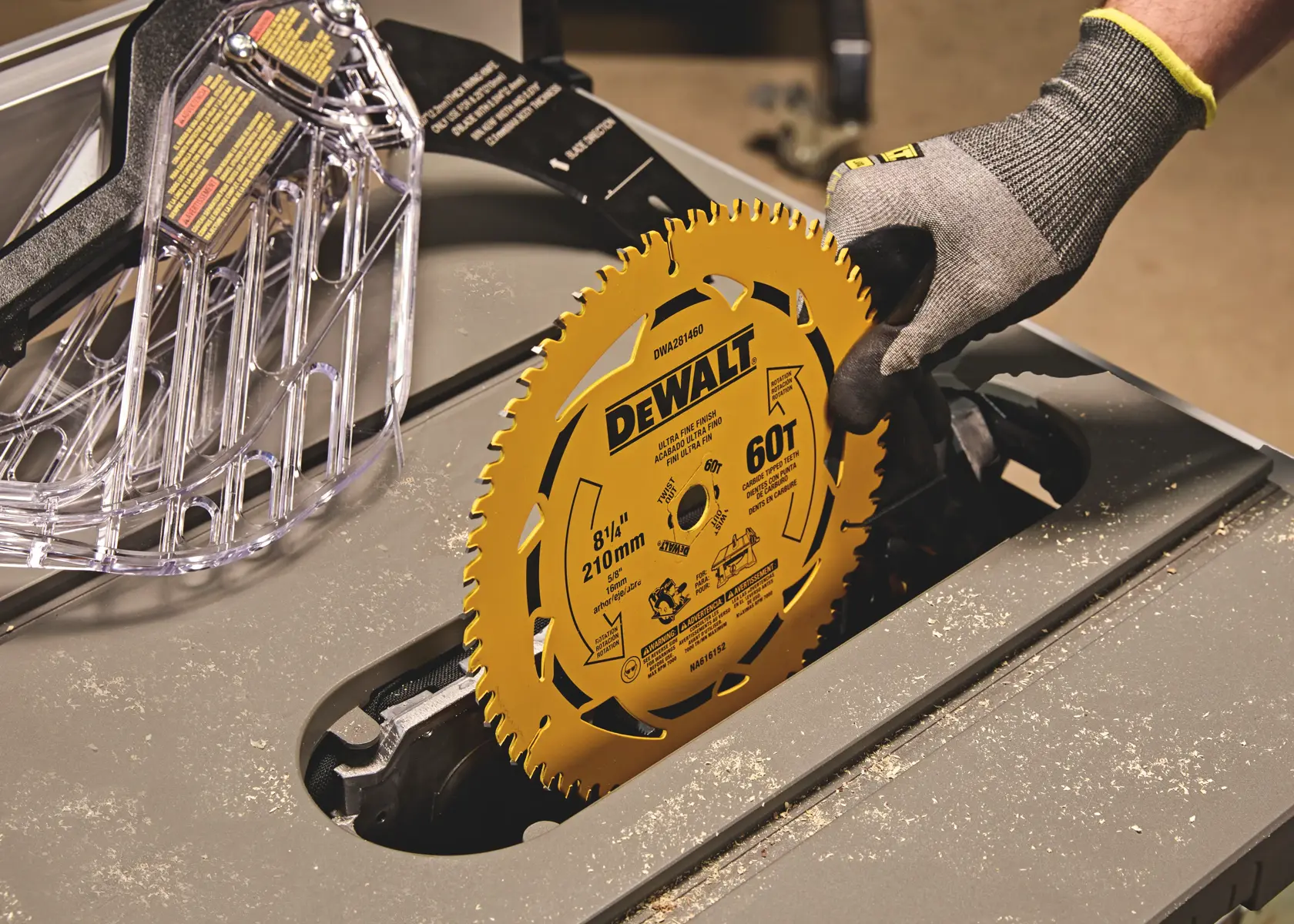 A gloved hand installing a yellow DEWALT circular saw blade, model DWA281460, onto a table saw. The blade is marked 8-1/4 inch (210 mm) and 60 teeth. The surrounding area shows sawdust and a transparent blade guard.