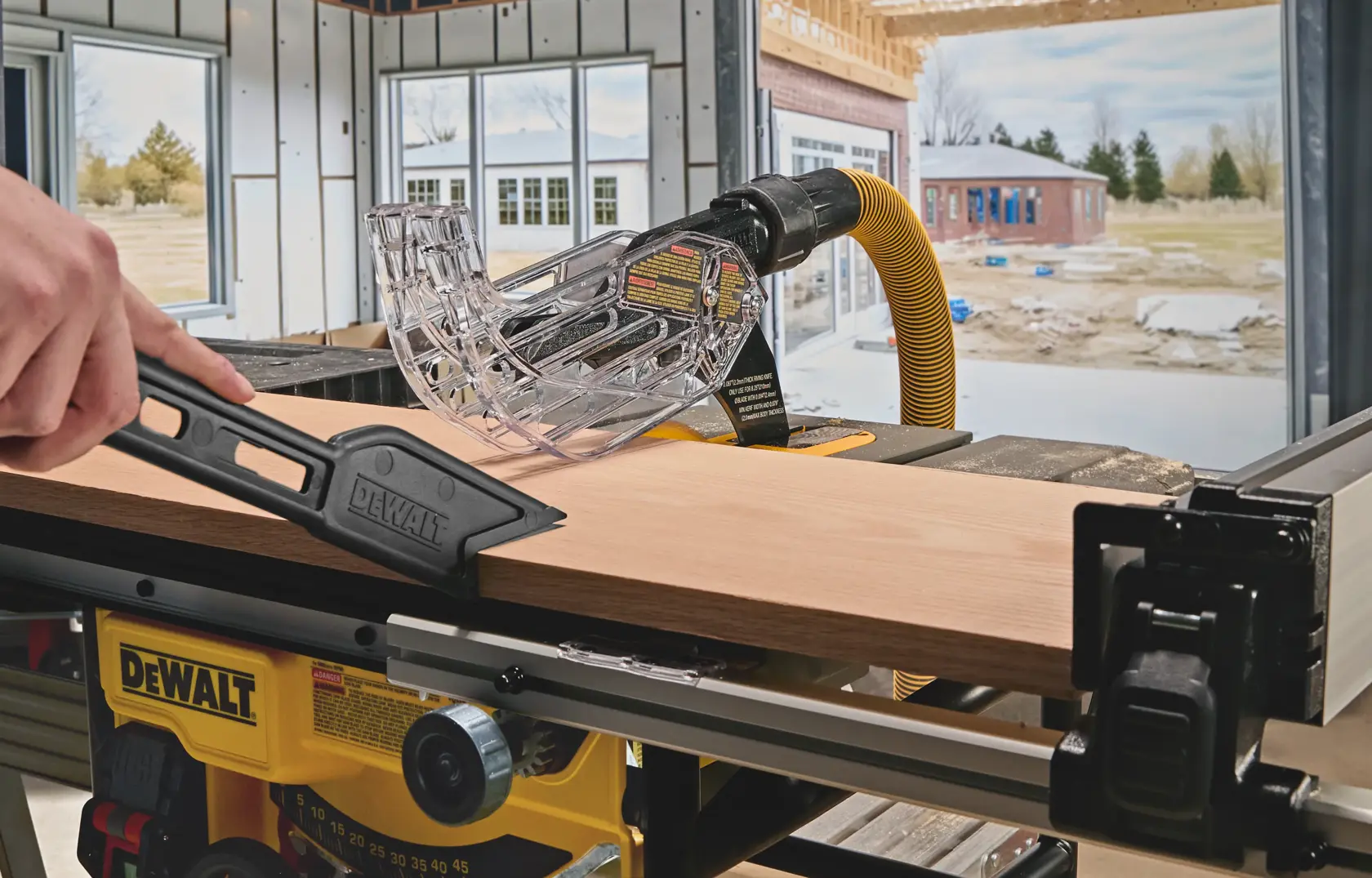 A DEWALT table saw in use, showing a hand guiding a piece of wood with a push stick under a clear blade guard, inside a partially constructed building with a window view of another house and some trees outside.
